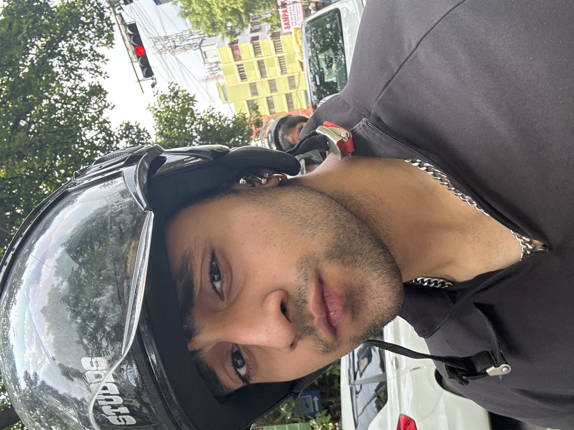 A man wearing a black helmet and a black jacket with a silver chain around his neck is in Kolkata, India. The traffic light shows red. Cars are parked in the background.