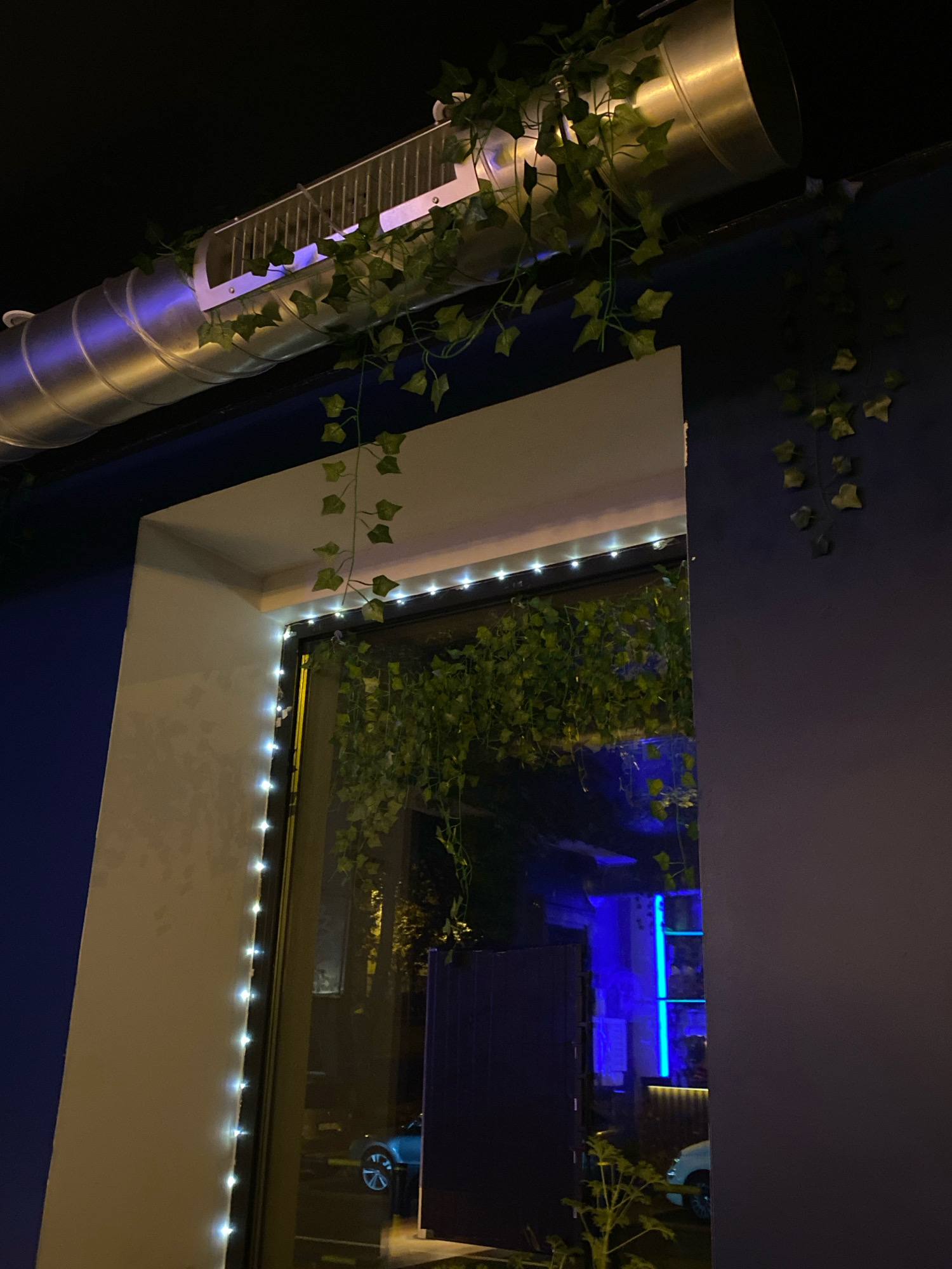The image depicts an outdoor nighttime scene with a focus on the facade of a building. A large, silver ventilation pipe runs horizontally across the top of the frame, adorned with artificial ivy. Below this, a doorway or large window is visible, framed by a white structure. A string of white fairy lights outlines the edge of this opening. Through the opening, the interior of a building is dimly lit, revealing blue neon lighting and some reflections of cars. The exterior walls of the building are painted in dark blue and a light, textured beige. The image is taken from a low angle looking upwards. This scene is located in the city of Gliwice, Poland.