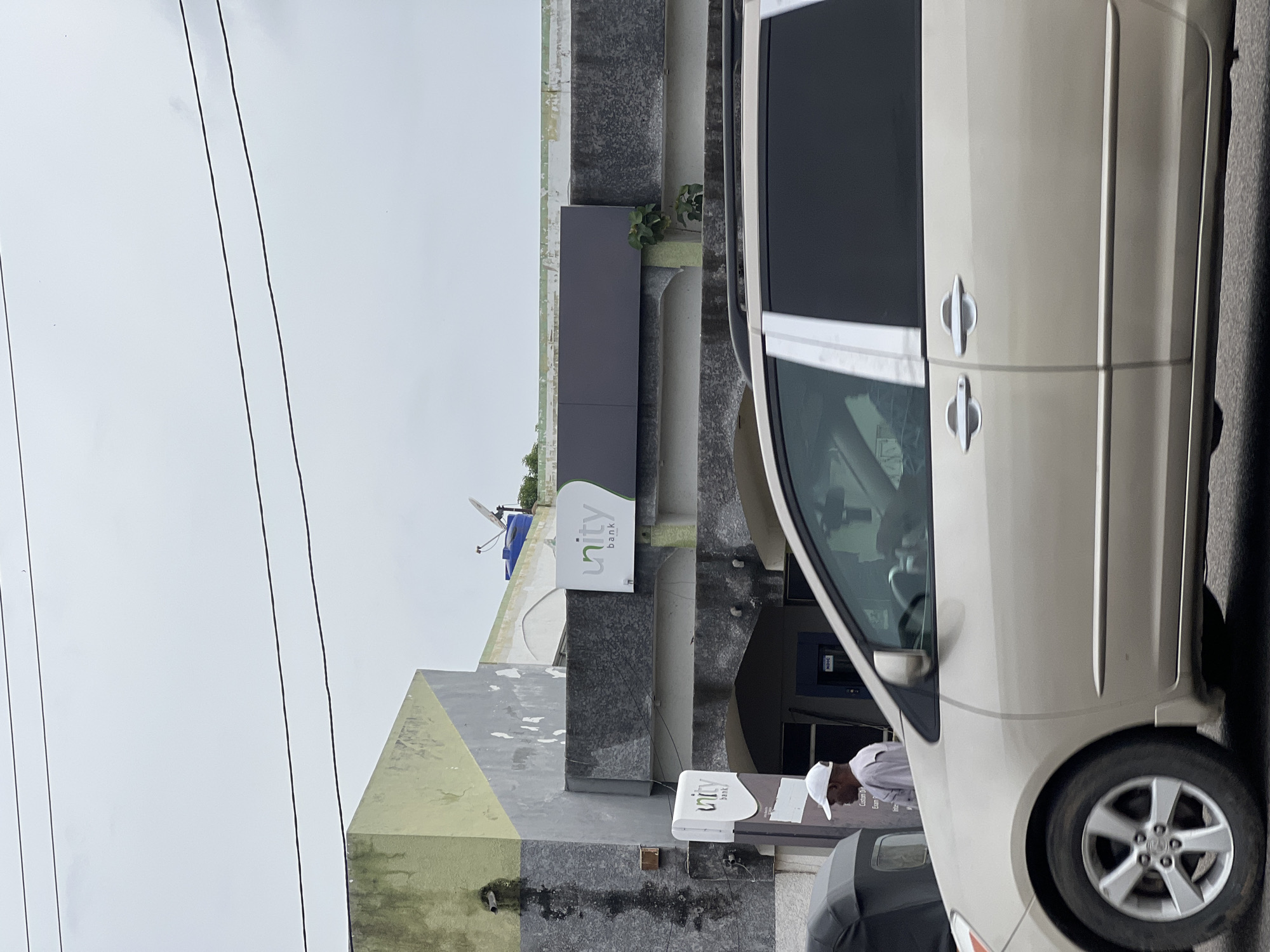 The image shows a street view in Jalingo, Nigeria. In the foreground, a beige minivan is parked on the right side of the frame. The minivan is partially obscured by the edge of the image on the left. The van has silver trim around the windows and door handles. On the left side of the frame, a person wearing a white cap and a grey long-sleeved shirt is standing near an ATM. To the left of the ATM, a dark-colored tricycle or motorcycle is visible.
Behind the ATM and the standing person, a building with a light green and grey facade is visible. The building has a sign with the "unity bank" logo. A darker rectangular sign is mounted above the "unity bank" sign. Further up on the building's roof, a blue water tank and a satellite dish are present. The sky is overcast and grey. The building appears to be in an urban environment, likely a commercial area.
