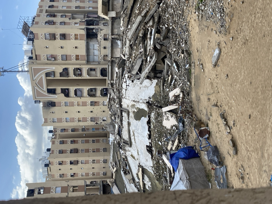 A destroyed building lies amidst rubble and debris, next to a taller, relatively intact apartment building. Damage is evident on the surviving structure with visible holes. The surrounding area is covered in concrete fragments and other remnants of destruction. A blue tarp and other makeshift coverings sit on the ground. The sky is blue with some white clouds.