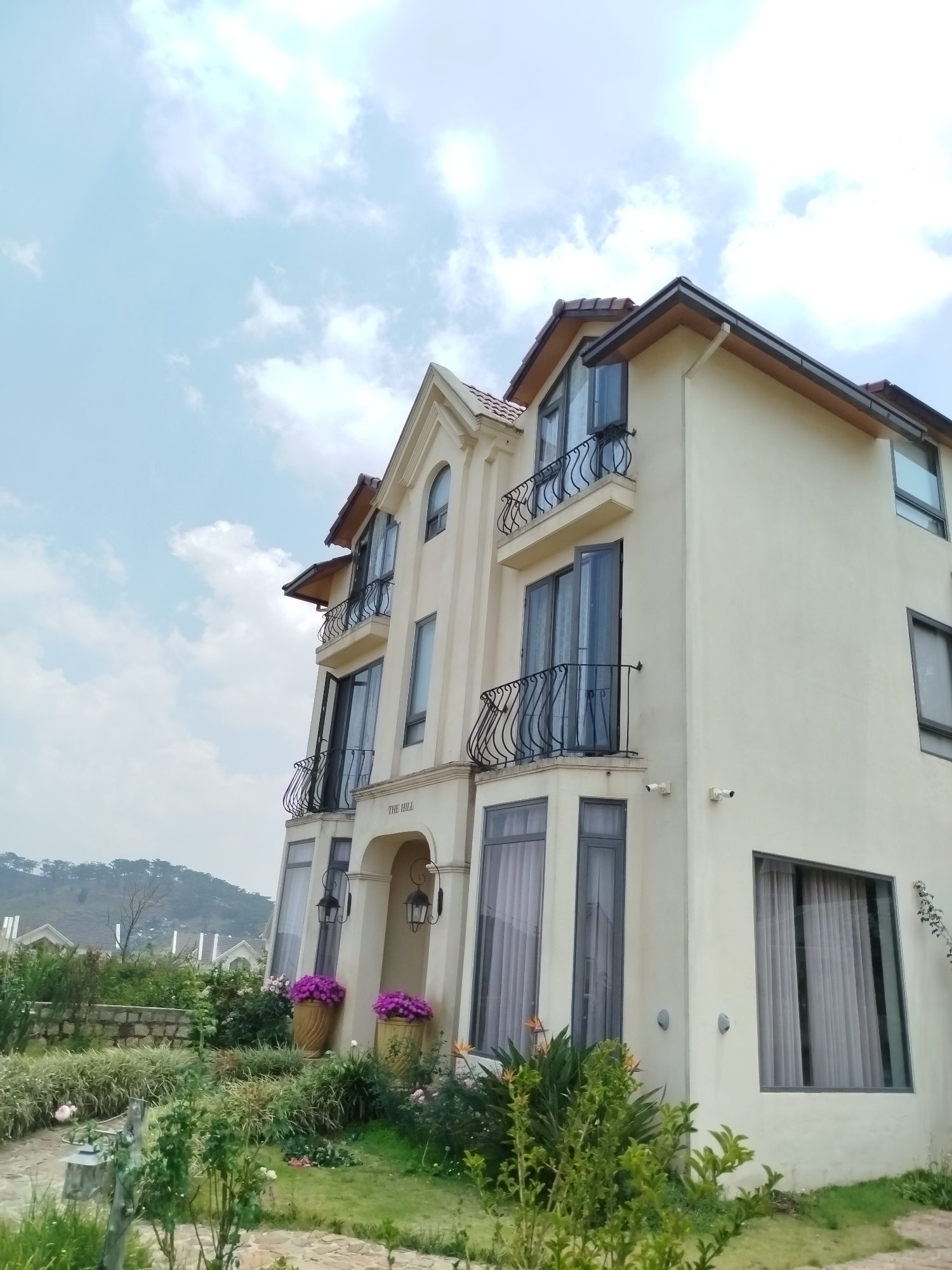 A three-story building with off-white stucco exterior walls is situated in Lang Biang - Đà Lạt, Vietnam. The building features arched entryways, wrought iron balconies, and a brown tiled roof. The facade has tall windows with gray frames. A sign above the entryway reads "The Hill." Potted purple flowers flank the entrance. A well-maintained green lawn and garden surround the building, extending towards a low stone wall and a distant forested hillside. The sky is partly cloudy. Two security cameras are affixed to the exterior walls.