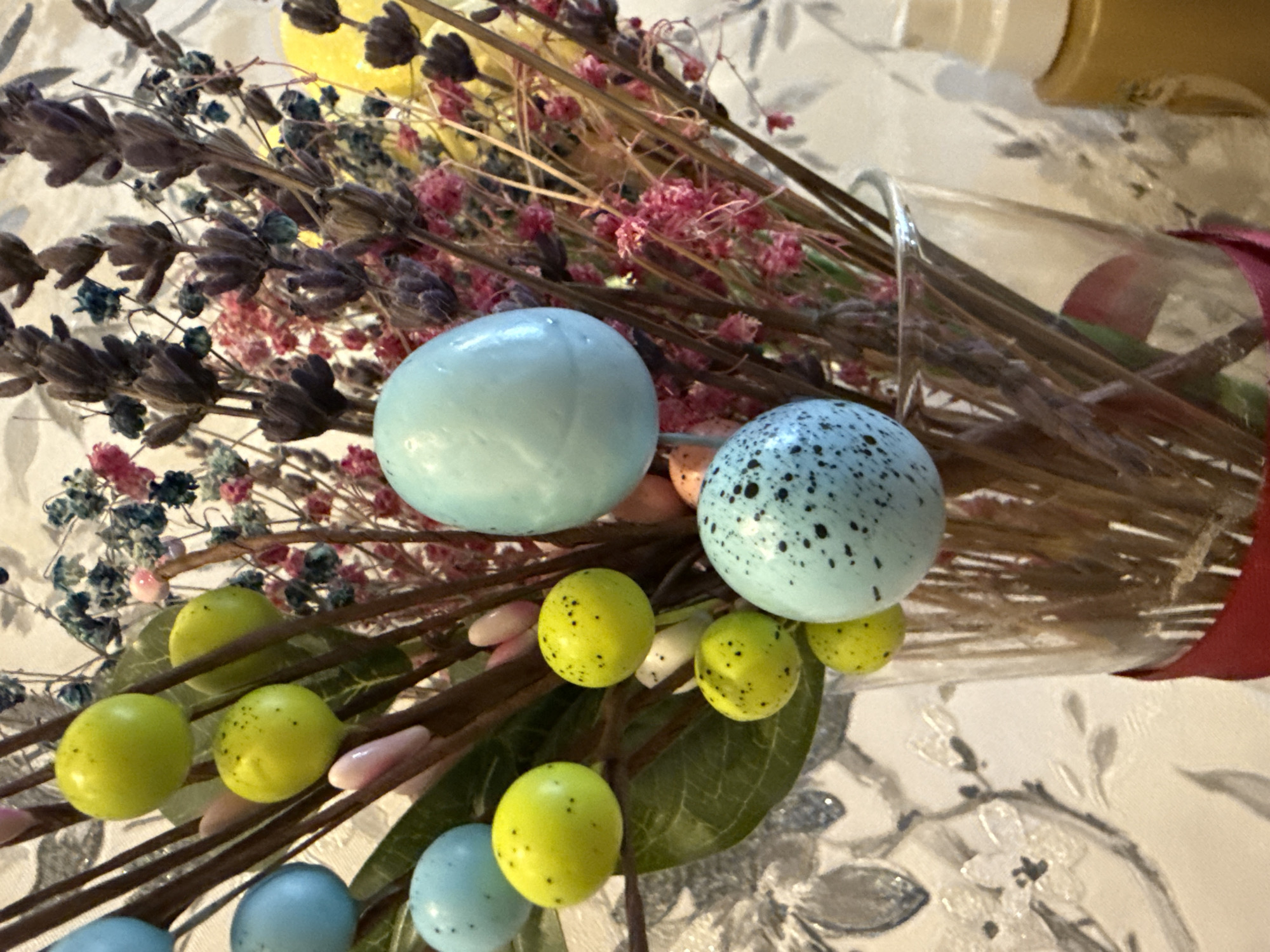 A close-up shot depicts a bouquet of dried flowers and decorative eggs, possibly an Easter arrangement. The flowers consist of lavender, pink, yellow, and blue blooms. The eggs are light blue and speckled. Yellow eggs are present as well. The bouquet is tied with a red ribbon and is in a clear vase. The background shows a decorative table. The location, Марковский, Russia, is irrelevant to the visual elements in the image.