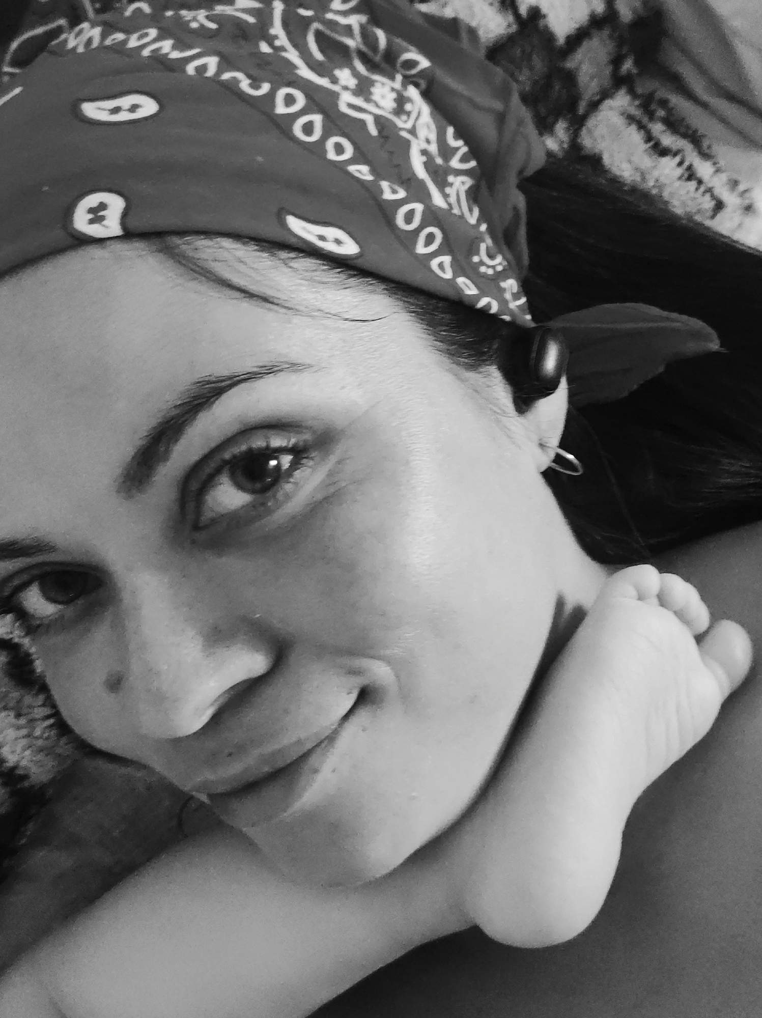 A close-up, black and white photograph features a woman wearing a bandana on her head, with a small child's foot resting near her neck. The woman is looking directly at the camera with a slight smile. A small earbud is visible in her ear. The background appears to be a textured fabric. No location is discernible from the image.