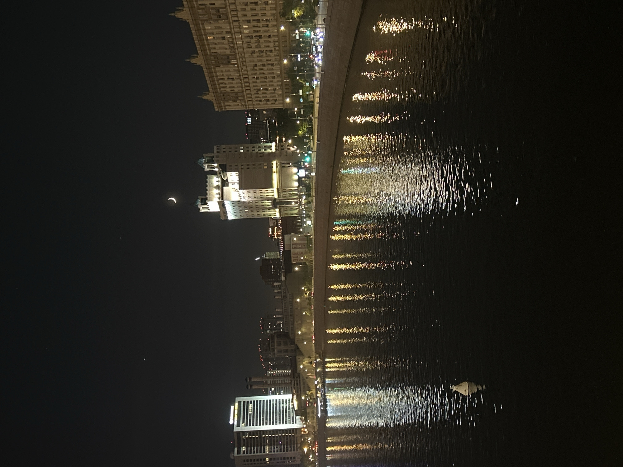 The image shows a nighttime cityscape reflected in a body of water, likely a river or large body of water. Along the riverbank, there are buildings of varying heights and architectural styles, illuminated by artificial lights. The reflections of these lights create shimmering patterns on the water's surface. In the dark sky, a crescent moon is visible. The location appears to be a city with a waterfront area.