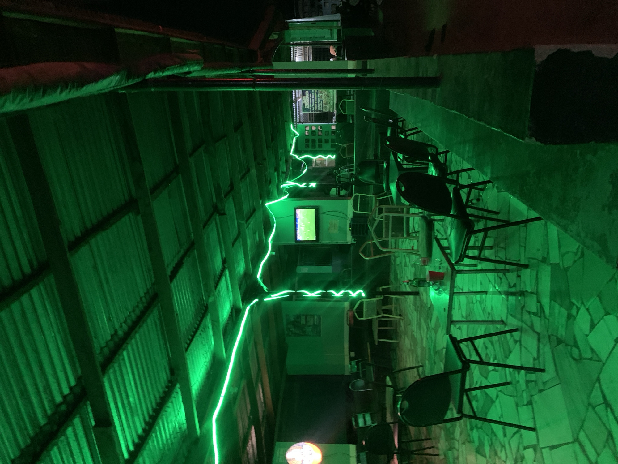 The image, rotated 90 degrees clockwise, depicts a long, narrow indoor or semi-enclosed space, likely a bar or casual restaurant, dominated by intense green lighting. A continuous, winding green LED light strip runs along the upper part of the left wall, which is constructed from corrugated metal paneling. The floor is made of irregularly shaped, light-colored stone tiles, entirely cast in green by the pervasive illumination.
On the right side of the space, multiple dark-colored chairs and tables are arranged along a wall, with additional lighter-colored chairs visible further into the background. The area appears set up for patrons but is currently devoid of people or activity. In the mid-ground, a television screen displays a green-tinged image.
Below the television, a sign is clearly visible. At the top, the text "Kuntop" can be read. Below this, a menu is listed, including beverages such as "Beer" (specifically mentioning "Star" and "Heineken"), "Malta Guinness," "Maltina," "Big Cola," "Fanta," "Sprite," and "Water." Food items listed include "Indomie noodles," "Fried Egg," "Bread," "Pepper Soup," "Isiewu," "Nkwobi," "Shawarma," and "Chicken."
The heavy artificial lighting suggests the scene takes place at night or in a perpetually dimly lit interior. The overall aesthetic is functional yet vibrant due to the monochrome green light. The scene appears to be an establishment ready for business, located in a context such as New Stadium, Nigeria.