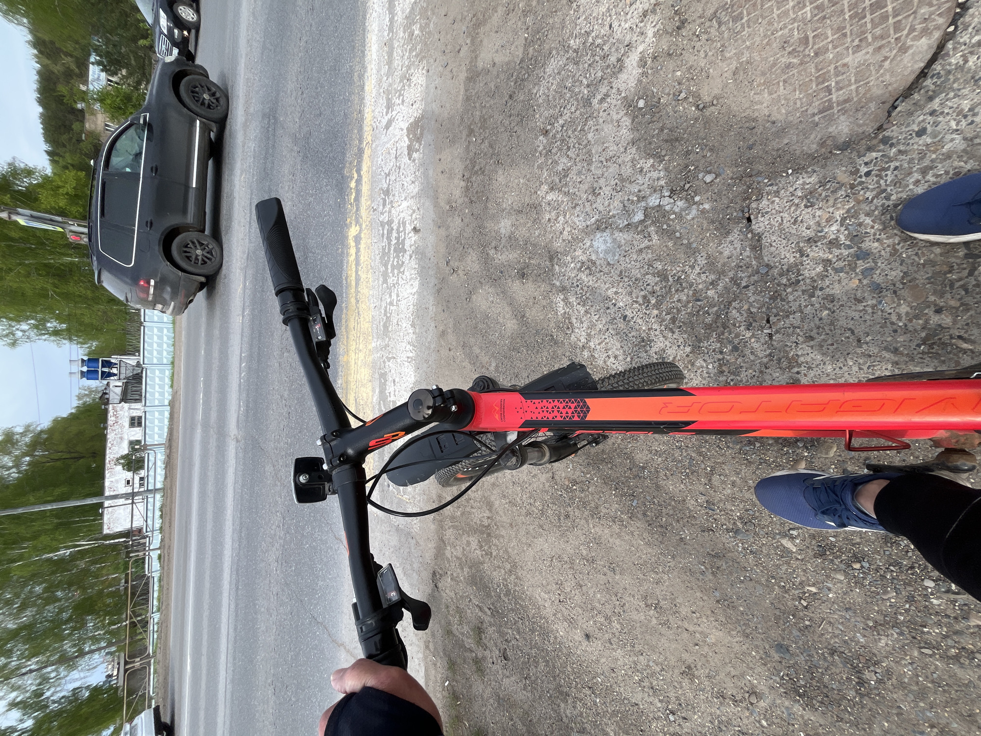 A first-person, point-of-view image shows a person riding a red and black "VICTOR" mountain bike on the shoulder of a paved road. The person is wearing dark leggings and blue sneakers. Part of a dark SUV is visible on the road to the left. Trees and buildings are also visible on the left side of the road. The shoulder is gravel and concrete. The location is Chaikovsky, Russia.