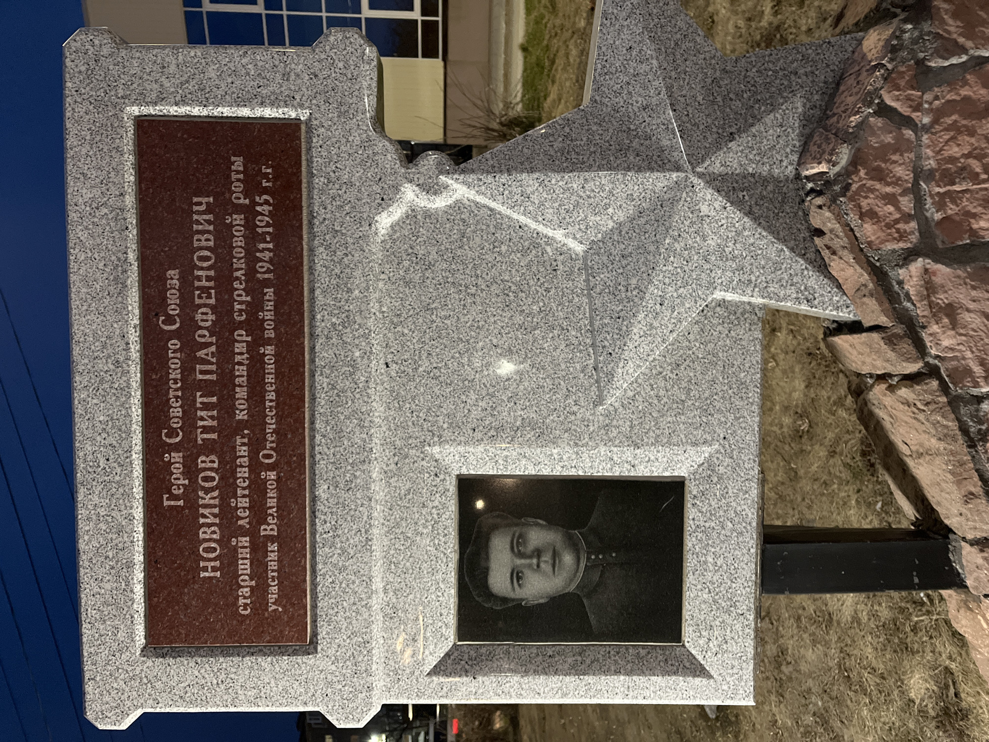 The image shows a memorial in Petropavlovsk-Kamchatsky, Russia, dedicated to Novikov Tit Parfenovich, a Hero of the Soviet Union. The memorial is made of gray granite and features a five-pointed star at the top. Below the star, there's a rectangular plaque with an inscription in Russian identifying Novikov Tit Parfenovich as a senior lieutenant and commander of a rifle company who participated in the Great Patriotic War from 1941 to 1945. There is a portrait of the man shown on the memorial. The memorial is positioned next to a small stone wall and some dry grass. In the background, there is a building and the sky is turning dark.