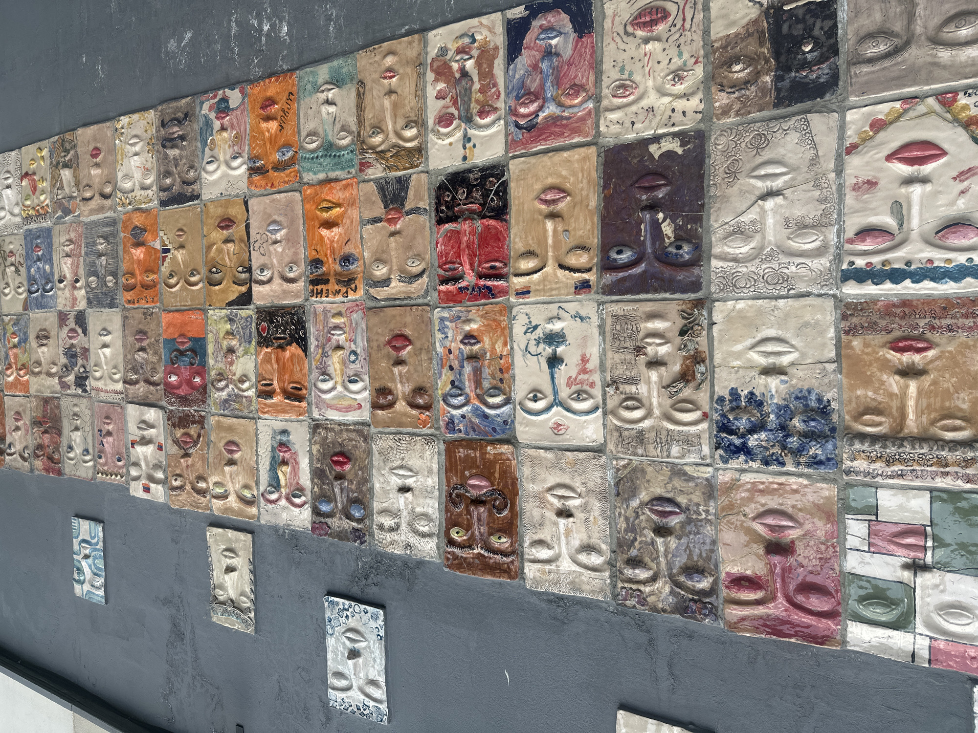 The image depicts a wall in Yerevan, Armenia, adorned with numerous ceramic tiles. Each tile features a stylized, raised relief face, painted in a variety of colors and patterns. The tiles are arranged in a grid-like pattern, covering a significant portion of the grey wall. Some tiles are mounted independently. The artwork appears to be a public art installation.