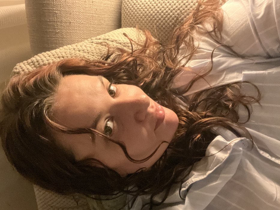 A woman with dark, curly hair and light skin is lying on a beige textured cushion or pillow. Her head is tilted, and she looks at the camera with wide eyes. She is wearing a light blue and white striped button-down shirt. The lighting is soft and warm, casting shadows across her face and hair.