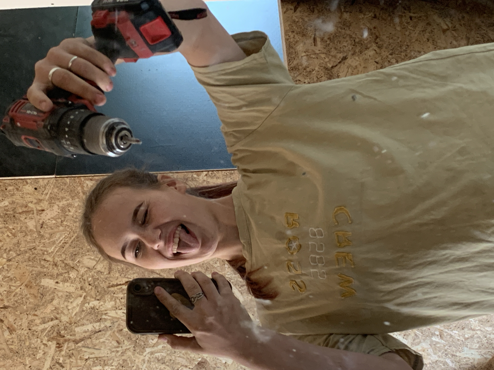 The image shows a person reflected in a mirror, holding a drill and a smartphone. The person is fair-skinned with light brown hair pulled back, and is wearing a light brown t-shirt with the word "CREW" and the number "2828" embroidered on it. They are sticking their tongue out and appear to be taking a selfie. The person has rings on their fingers on both hands. The drill is red and black. The background appears to be unfinished wood paneling and a blue surface. The location of Фоки, Russia, is not discernible from the image.