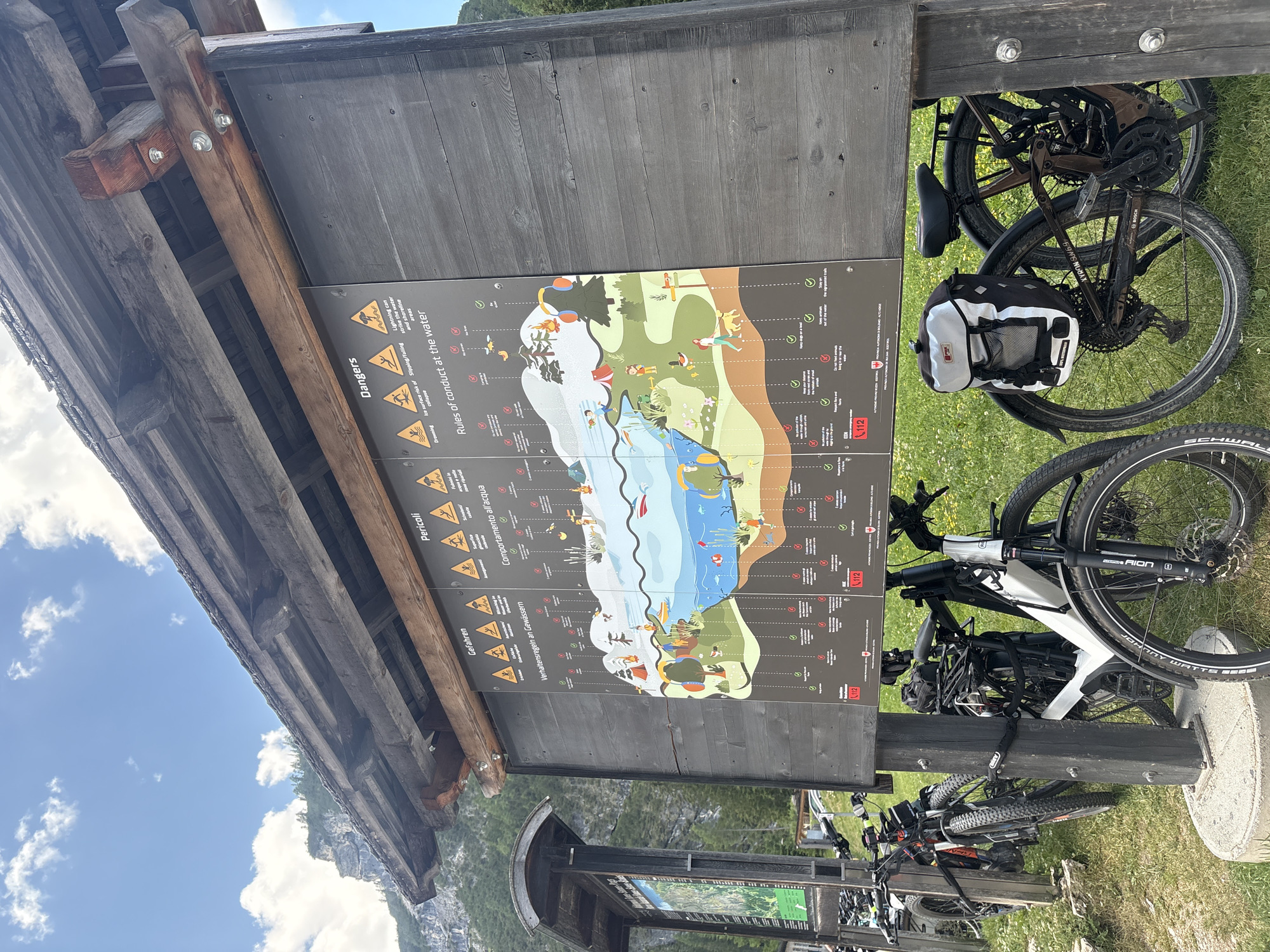 A wooden signpost with informational graphics and text is situated outdoors in a grassy area. The sign has a dark wooden background with a wooden overhang. On the sign, there are three columns of text in German, Italian, and English, respectively, along with illustrations depicting potential dangers and rules of conduct at the water. The German column is titled "Gefahren" and "Verhaltensregeln an Gewässern." The Italian column is titled "Pericoli" and "Comportamento all'acqua." The English column is titled "Dangers" and "Rules of conduct at the water." Several bicycles are parked in front of the signpost, chained together. One bicycle is white with black accents, another is black and orange, and a third is dark brown. The environment appears to be mountainous, with trees and a cloudy sky visible in the background. The ground is covered in green grass and scattered small stones.