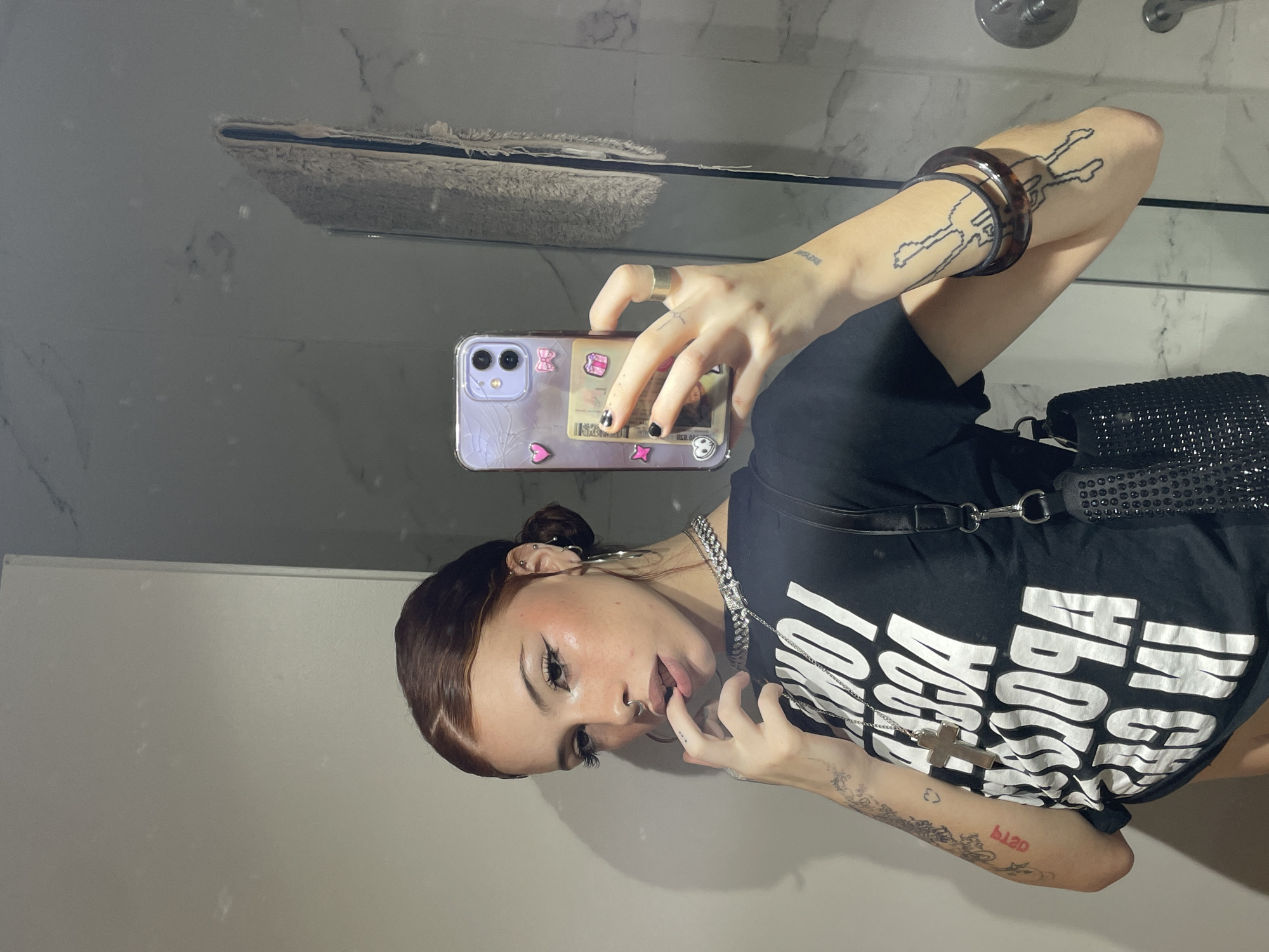 A person is taking a selfie in a bathroom. The person has light skin, brown hair pulled back in a bun, and multiple ear piercings. They are wearing a black t-shirt with white text, a silver chain necklace, and bracelets on their left arm. The phone case is clear with stickers and a cracked design. The bathroom has white walls and a mirror. There is a shower head visible in the background, and a towel hanging on the wall. There is no indication of the city of Buenos Aires, Argentina.