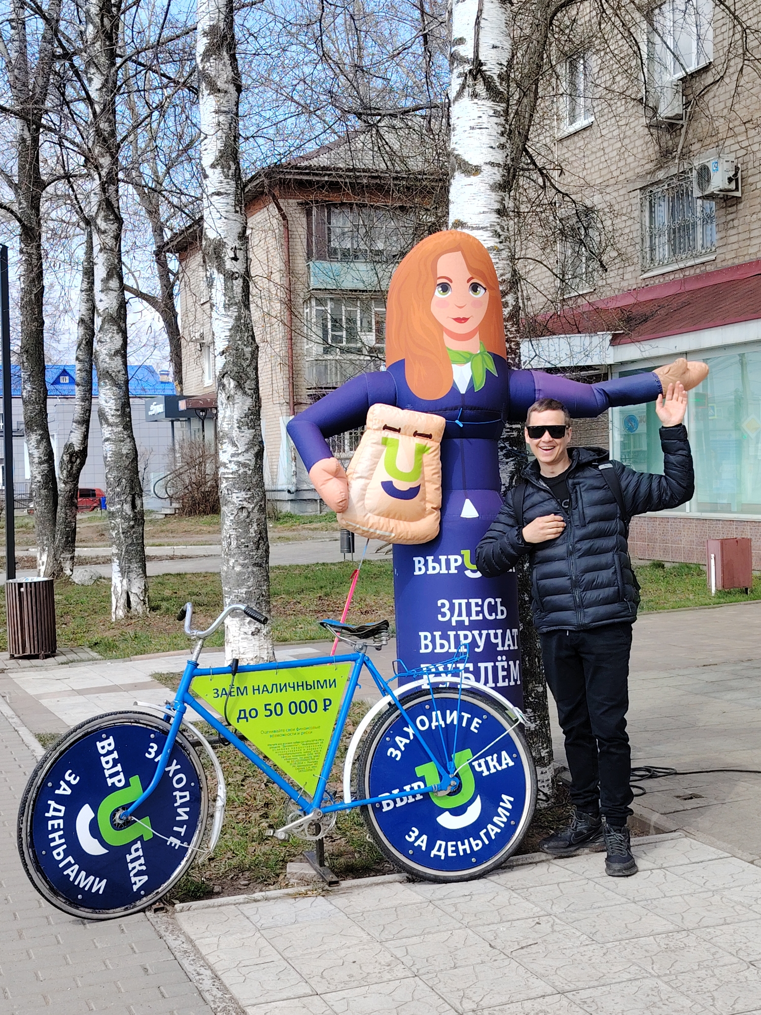 The image captures a bright, daytime street scene in Chaikovsky, Russia, likely in early spring given the bare branches of the trees. A smiling man, wearing a black puffer jacket, sunglasses, and a backpack, stands on a paved sidewalk, posing for the camera. He gestures with one hand raised and the other on his chest, appearing cheerful.
He is positioned next to a large, purple inflatable advertising figure depicting a cartoon woman with red hair, a green scarf, and a blue suit, holding a light brown bag with a green 'U' logo. The base of the inflatable reads in Russian: "ЗДЕСЬ ВЫРУЧАТ РУБЛЁМ" (Here they will help with rubles), "ЗАХОДИТЕ" (Come in), "ВЫРУЧКА" (Revenue/Help out), and "ЗА ДЕНЬГАМИ" (For money), accompanied by the green 'U' logo.
Adjacent to the man and inflatable is a blue bicycle, also used for advertisement. A green sign attached to its frame states: "ЗАЁМ НАЛИЧНЫМИ" (Cash Loan) "до 50 000 Р" (up to 50 000 R), along with "Оценивайте свои финансовые возможности и риски" (Assess your financial capabilities and risks). The bicycle's wheels are covered with blue discs featuring the same "ЗАХОДИТЕ", "ВЫРУЧКА", "ЗА ДЕНЬГАМИ" text and the green 'U' logo, reinforcing the promotional message for financial services.
Multiple tall birch trees line the sidewalk, and in the background, there is a light-colored brick building with windows, some having security bars, and a red roof. Another building with a blue roof is visible further away. A dark brown trash can stands on the left side of the frame.