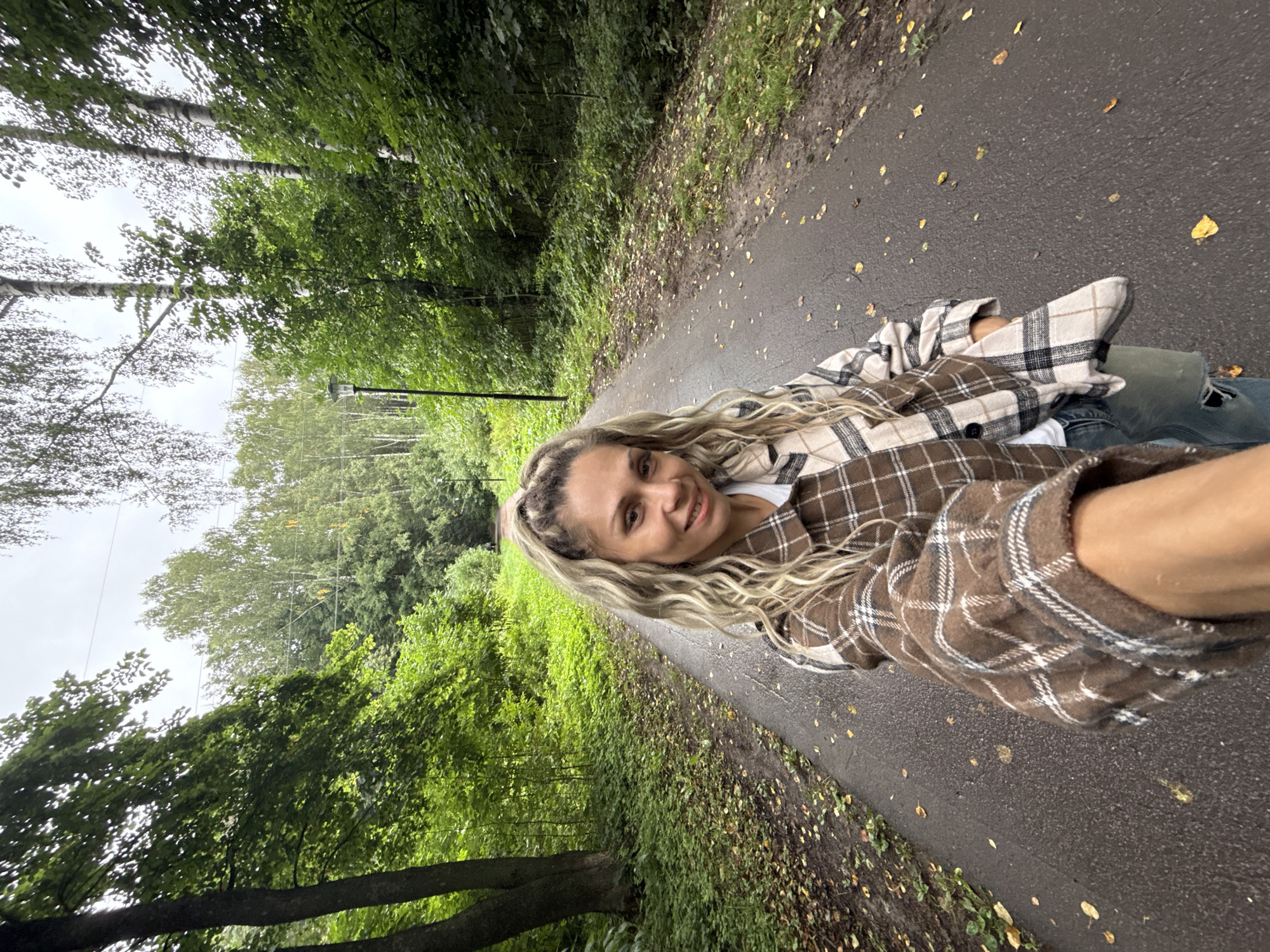 A medium shot captures a woman with long, blonde, wavy hair tied in a ponytail from a selfie perspective. She is wearing a brown and white plaid overshirt over a white t-shirt and appears to be standing on a paved path in a park. The path is flanked by lush green trees and foliage, with fallen leaves scattered on the ground. The sky is overcast. A lamppost is visible in the background. The scene appears to be in a park or natural setting.