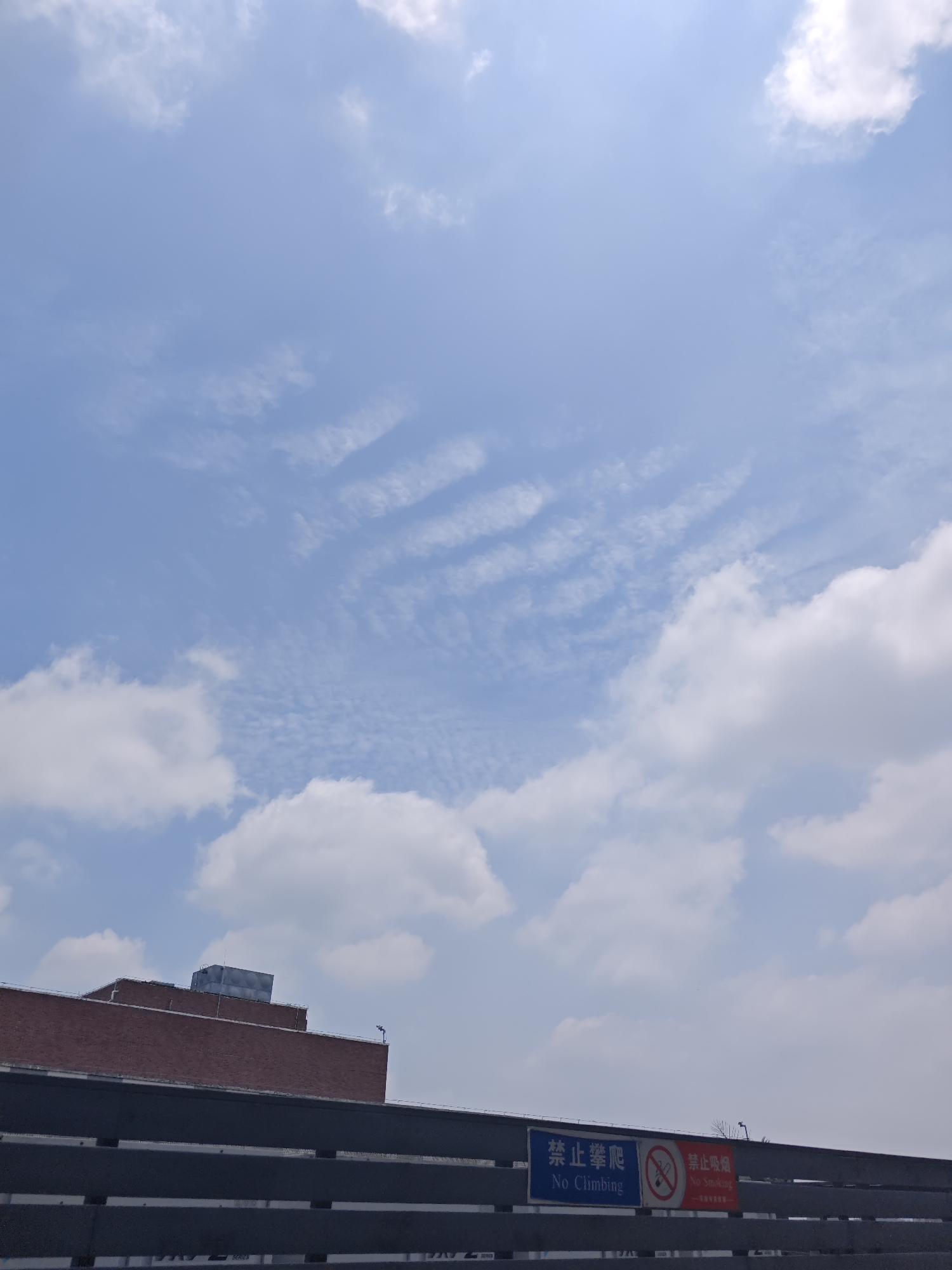 In the Chongqing High-tech Zone, China, the sky is a vibrant blue with scattered white clouds, some appearing wispy and others fluffy. In the lower portion of the image, a brick building with an industrial air conditioning unit on its roof is visible. Below the building, a dark grey structure runs horizontally across the frame, featuring two signs. The sign on the left is blue with white text and a prohibition symbol, reading "禁止攀爬" and "No Climbing". The sign on the right is red with white text and a prohibition symbol, reading "禁止吸烟" and "No Smoking".