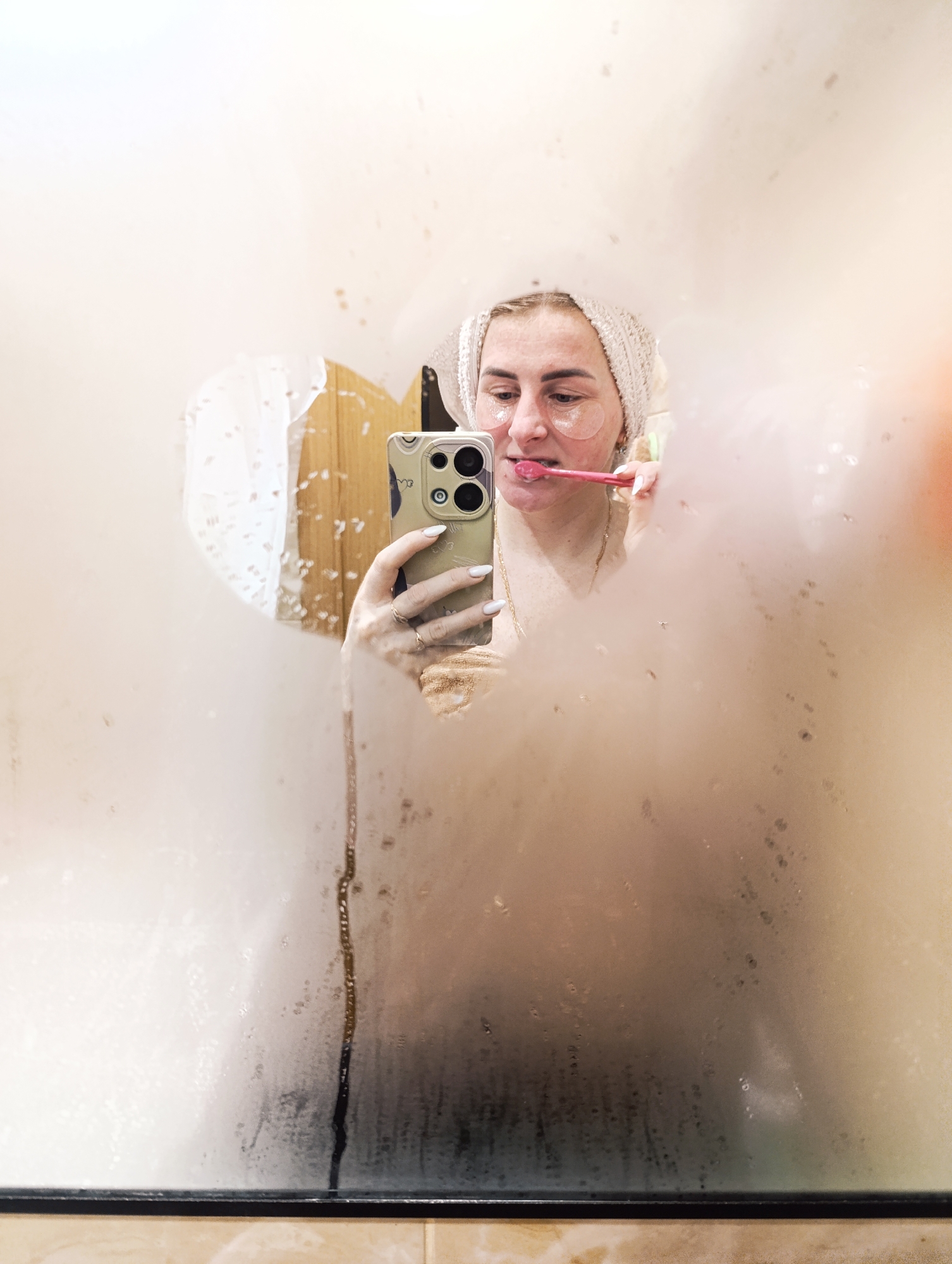 A young woman is seen in the reflection of a fogged-up bathroom mirror in an indoor setting. She wears a white towel on her head and eye patches, and is holding a pink toothbrush in her mouth while holding a smartphone in her right hand, apparently taking a selfie. The smartphone has a light green case. The mirror is partially wiped clear in a heart shape, and there are dark streaks of water running down the mirror's surface. The base of the mirror is mounted on a countertop with a dark trim and a cream-colored tile. The city and country are not discernible from the image.
