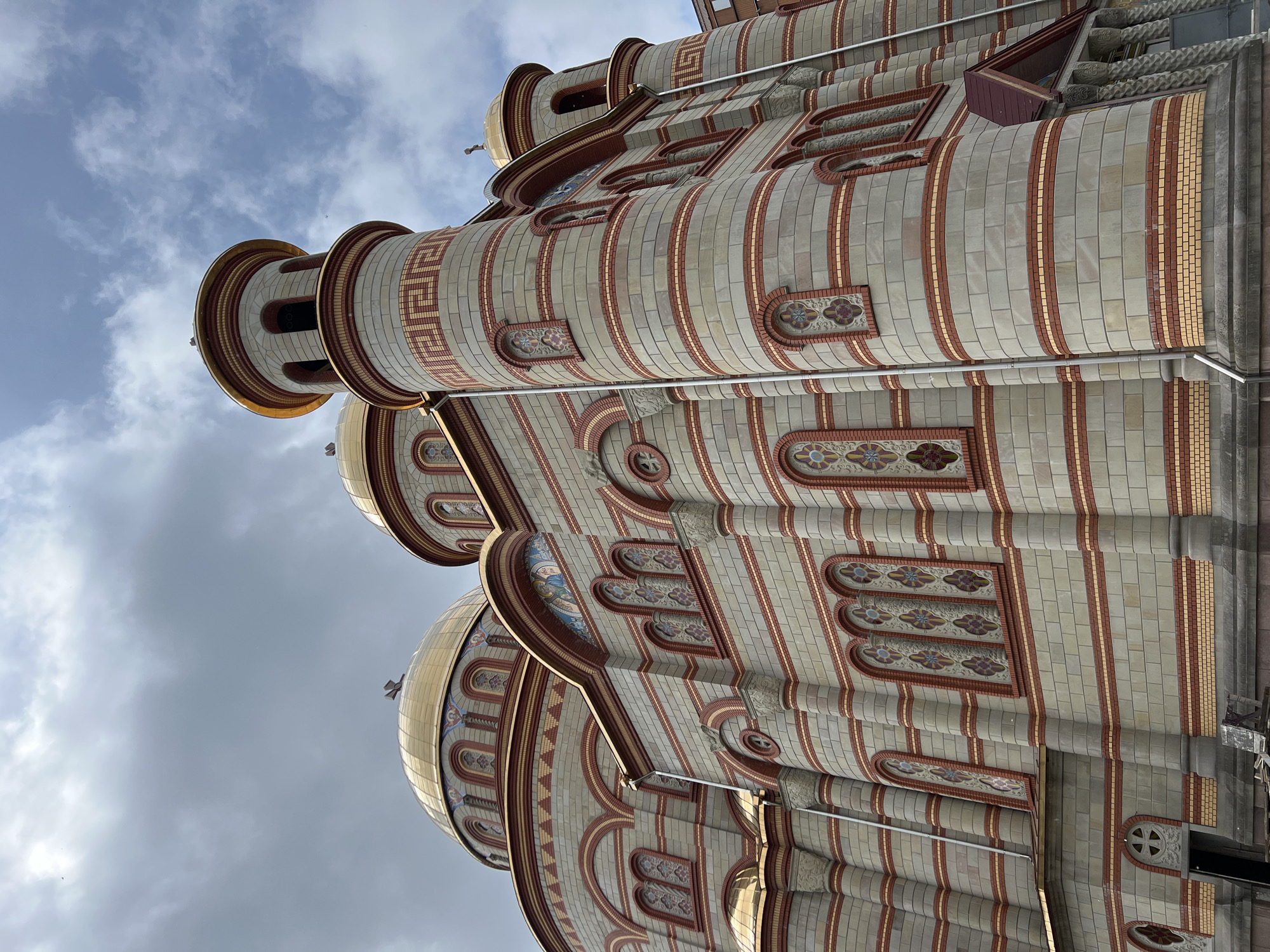 An upward-angled exterior shot captures the elaborate architecture of a large religious building, likely a cathedral or church, under an overcast sky in Ternopil, Ukraine. The structure features multiple golden domes, several of which are visible, each topped with a cross. The facade is predominantly constructed from light grey or cream-colored bricks, interspersed with decorative vertical and horizontal bands of red and yellow bricks.
The building's design incorporates numerous arched windows and decorative elements. Some windows are tall and narrow, while others are small and circular, often featuring intricate patterns or stained-glass details. Above many of the arches, colorful mosaic-like panels depict figures or ornate floral motifs. Cylindrical towers with small, arched openings rise from the structure, adorned with geometric patterns, including a prominent Greek key design.
The overall impression is one of grandeur and intricate craftsmanship, characteristic of Orthodox Christian architecture. No people or activities are visible, emphasizing the building itself. The cloudy sky adds a dramatic backdrop to the ornate structure, suggesting daytime with diffuse natural light. No discernible text is visible on the building.