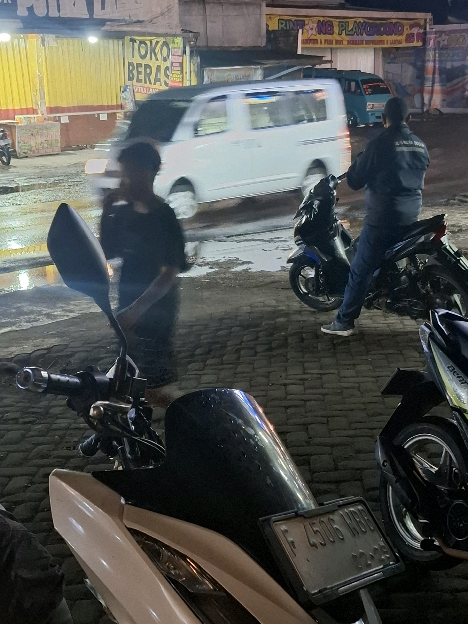 The image depicts a street scene at night, likely in Ciheulang Tonggoh, Indonesia, with several motorcycles and vehicles present. A person in a dark jacket and jeans is seated on a dark blue motorcycle in the mid-ground, facing away from the camera. To the left, another individual wearing a dark t-shirt and shorts stands near a motorcycle, with their head turned and holding a phone to their ear. In the background, a white van is moving, and a small blue car is parked. Buildings with illuminated signs are visible, including one that reads "TOKO BERAS" (Rice Shop) and another with "RINTING PLAYGROUND" and "MAINTENANCE & FREE WIFI". The ground is paved with cobblestones, and there are reflections on the wet surface, suggesting recent rain. A license plate on a motorcycle in the foreground reads "F 4506 WBB".