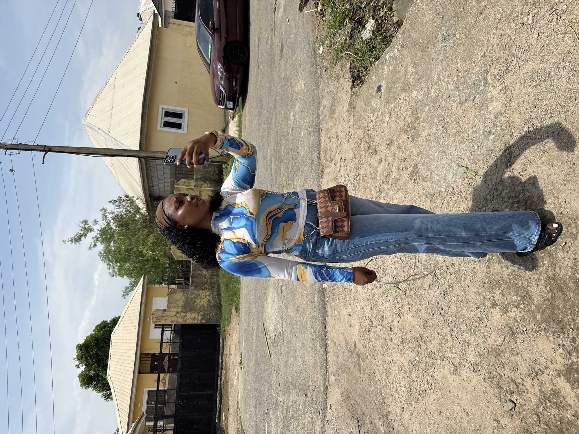 A vertical, full-body outdoor shot captures a young Black woman standing on a roughly paved or dirt road, holding a smartphone. She is wearing a long-sleeved top with a vibrant blue, white, and yellow abstract pattern, light blue jeans, and black sandals with white nail polish. A brown checkered crossbody bag with a gold chain strap hangs over her right hip. She has a brown headband or scarf and a gold watch on her left wrist. Her dark, curly hair is visible around her face.
The woman is holding a smartphone with a triple-lens camera setup (resembling an iPhone) in her right hand, raised as if taking a photo or video. In her left hand, she holds a white cable-like object, possibly a charging cable or earphones.
The background features residential buildings with yellow walls and corrugated metal roofs under a bright, sunny sky with scattered clouds. A dark red car is parked near one of the houses. Electrical poles and overhead wires crisscross the upper part of the image. Green trees and sparse vegetation are visible around the houses and along the roadside. The ground is a mix of concrete and dirt, with small stones and debris. Strong sunlight casts clear shadows, indicating it is daytime. The scene depicts a typical residential street, consistent with the provided context of Abuja, Nigeria. There is no visible text.