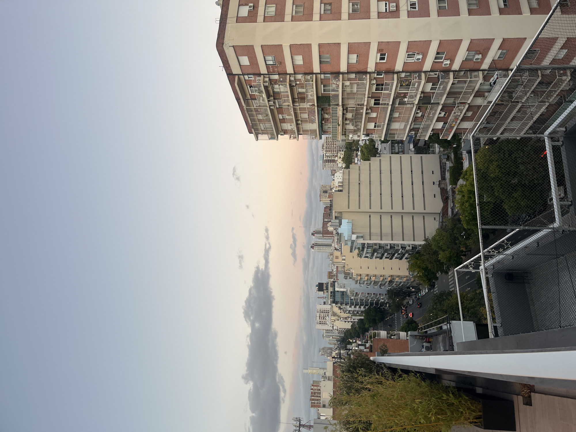 The image depicts a cityscape in Buenos Aires, Argentina, taken from a high vantage point, likely a rooftop. The foreground shows a rooftop with a safety fence and some greenery. In the middle ground, there are several buildings of varying heights and architectural styles. The buildings are mostly residential, with balconies and windows visible. A street runs between the buildings, with some cars visible. The sky in the background is a mix of light blue, gray, and pastel colors. The image has a slightly rotated perspective.