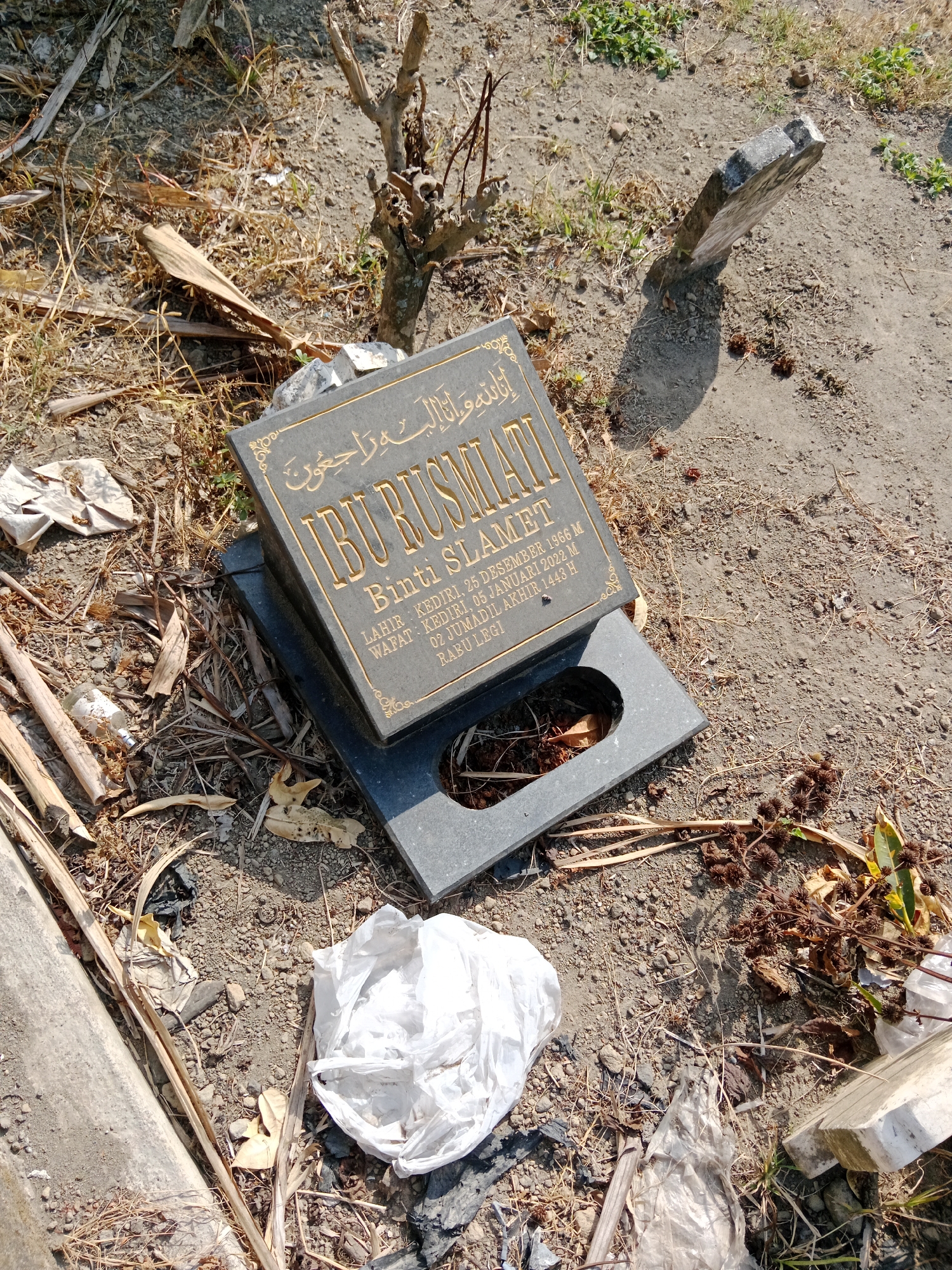 The image depicts a tombstone in what appears to be a graveyard in Susuhbango, Indonesia. The tombstone, made of dark stone with gold lettering, has inscriptions in both Arabic and Indonesian. The Arabic inscription at the top reads "إنا لله وإنا إليه راجعون" (Indeed we belong to Allah and indeed to Him we will return). Below this, the name "IBU RUSMANI" is prominently displayed. Further text on the tombstone indicates birth and death dates. "LAHIR: KEDIRI, 25 DESEMBER 1966 M" translates to "Born: Kediri, December 25, 1966". "WAFAT: KEDIRI, 05 JANUARI 2023 M" translates to "Died: Kediri, January 5, 2023". Below this, "2 DJUMADIL AKHIR 1443 H" is the Islamic calendar date of death. The tombstone is tilted and rests on a smaller base. The surrounding environment is unkempt, with dry grass, dead leaves, twigs, and scattered pieces of white plastic bags. In the background, a broken tree trunk stands, and another smaller, weathered stone is visible, leaning precariously. The ground is a mix of dry soil and gravel. The scene suggests neglect and possibly a less-maintained section of a cemetery.