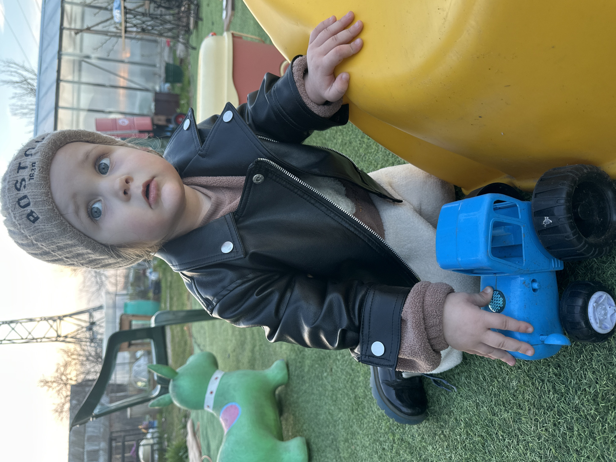 A toddler stands on artificial turf next to a yellow slide, holding a blue toy truck. The child is dressed in a brown knit hat, a black leather jacket, a brown shirt, and shiny black shoes. In the background, there is a greenhouse-like structure and a green dog-shaped object. The image was taken in Chaikovsky, Russia.