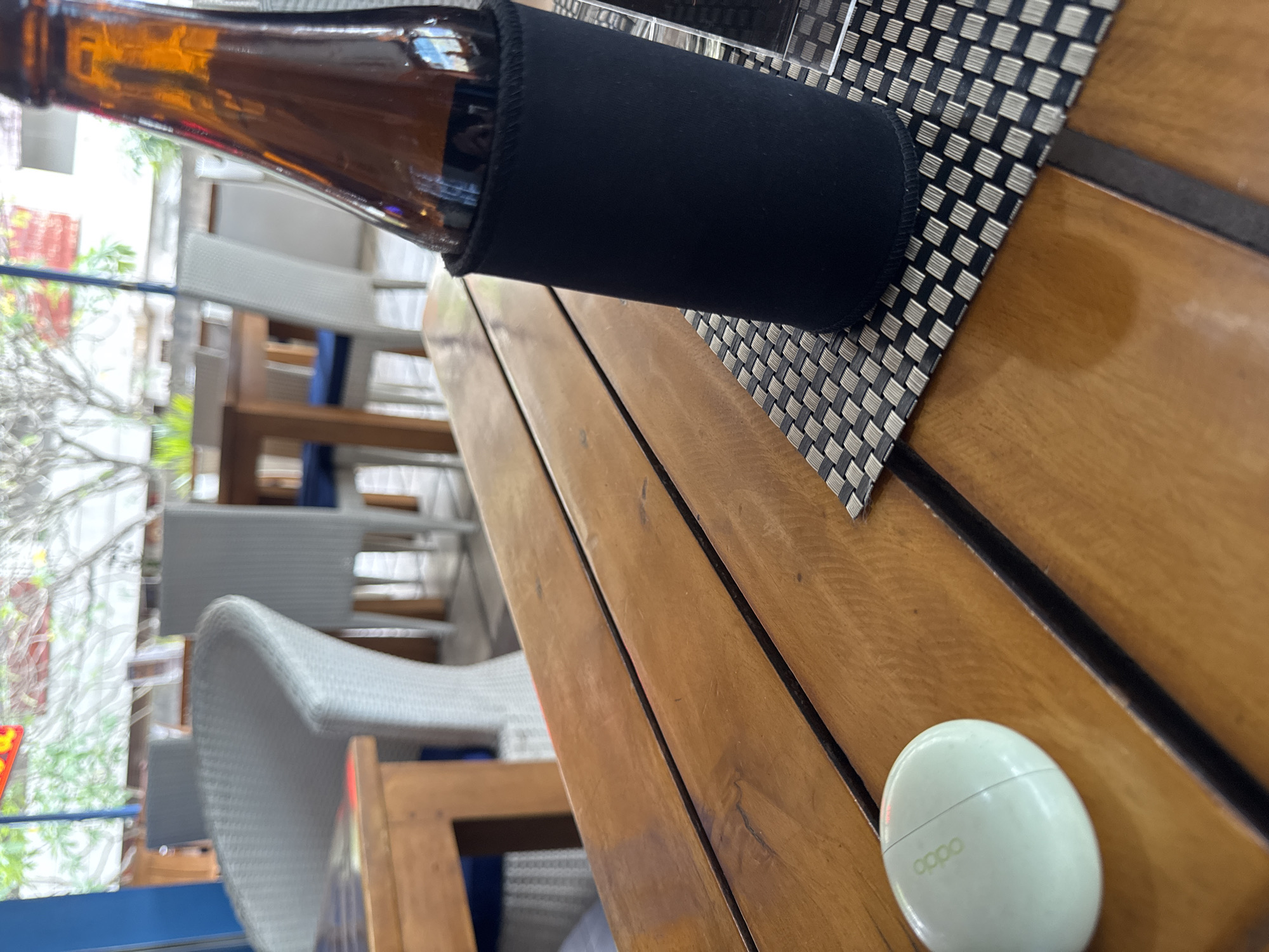 A close-up, eye-level shot focuses on a wooden table in an outdoor seating area at a restaurant in North Kuta, Indonesia. On the right side of the table, a dark brown glass bottle, partially covered by a black koozie, stands upright. Next to the bottle, a black and grey woven placemat is visible. To the left of the bottle and placemat, a light green oval-shaped object, likely a charging case for wireless earbuds, sits on the wooden table surface. The background is blurred, showing outdoor furniture, specifically white wicker chairs and wooden tables, suggesting a casual dining environment. The overall scene conveys a relaxed atmosphere, possibly a casual meal or drink at an establishment.