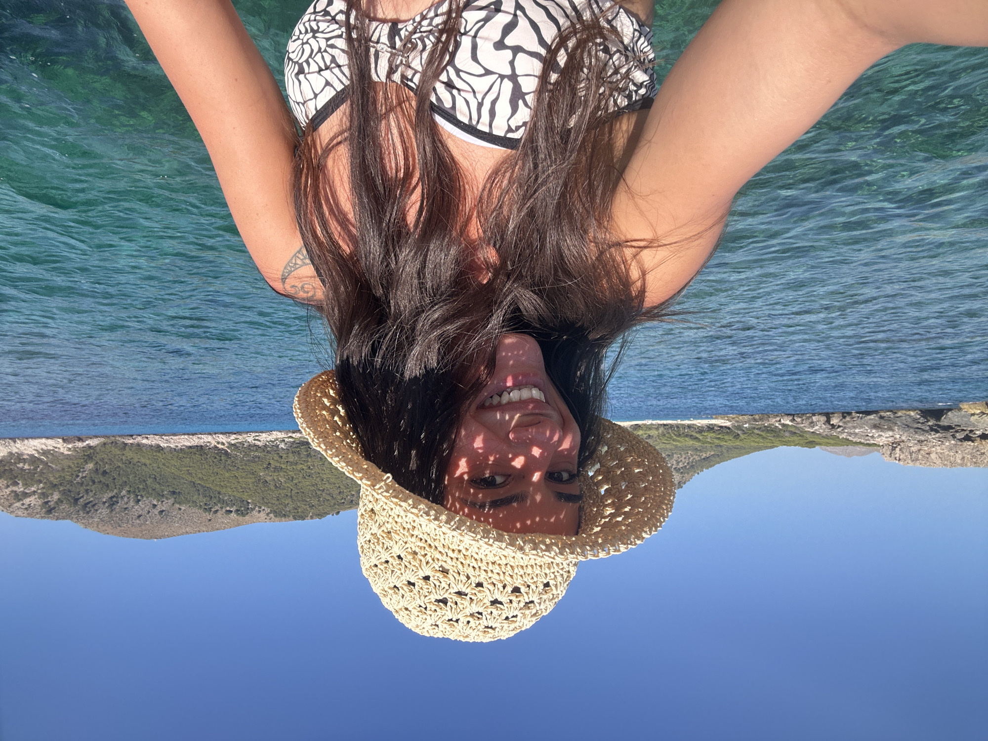 In Cala Lliteres, Spain, a woman with long, dark hair is captured in a selfie. She is wearing a straw hat that casts a patterned shadow on her face, and a black and white patterned bikini top. The clear, turquoise water of the sea surrounds her, with rocky shores and green, tree-covered hills visible in the background under a bright blue sky.