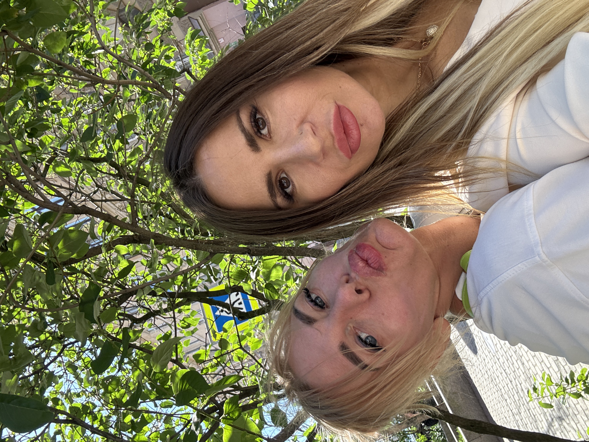 This is a selfie taken outdoors, possibly in Saint Petersburg, Russia, as indicated by the context. Two women are the primary subjects, positioned at the top and bottom of the frame. The woman at the top has long, brown and blonde hair, brown eyes, and is wearing a white top. The woman at the bottom has blonde hair, blue eyes, and is also wearing a white top with a green collar. Both women are looking towards the camera, with the lower woman making a kissing gesture.
They are positioned amongst the branches and green leaves of a tree, with sunlight filtering through the foliage. In the background, a building with windows is visible, and a blue and yellow sign with some lettering can be seen on the tree. The lighting suggests it is daytime, likely sunny. The overall mood appears casual and cheerful, typical of a selfie taken with friends. The image is rotated 90 degrees clockwise.