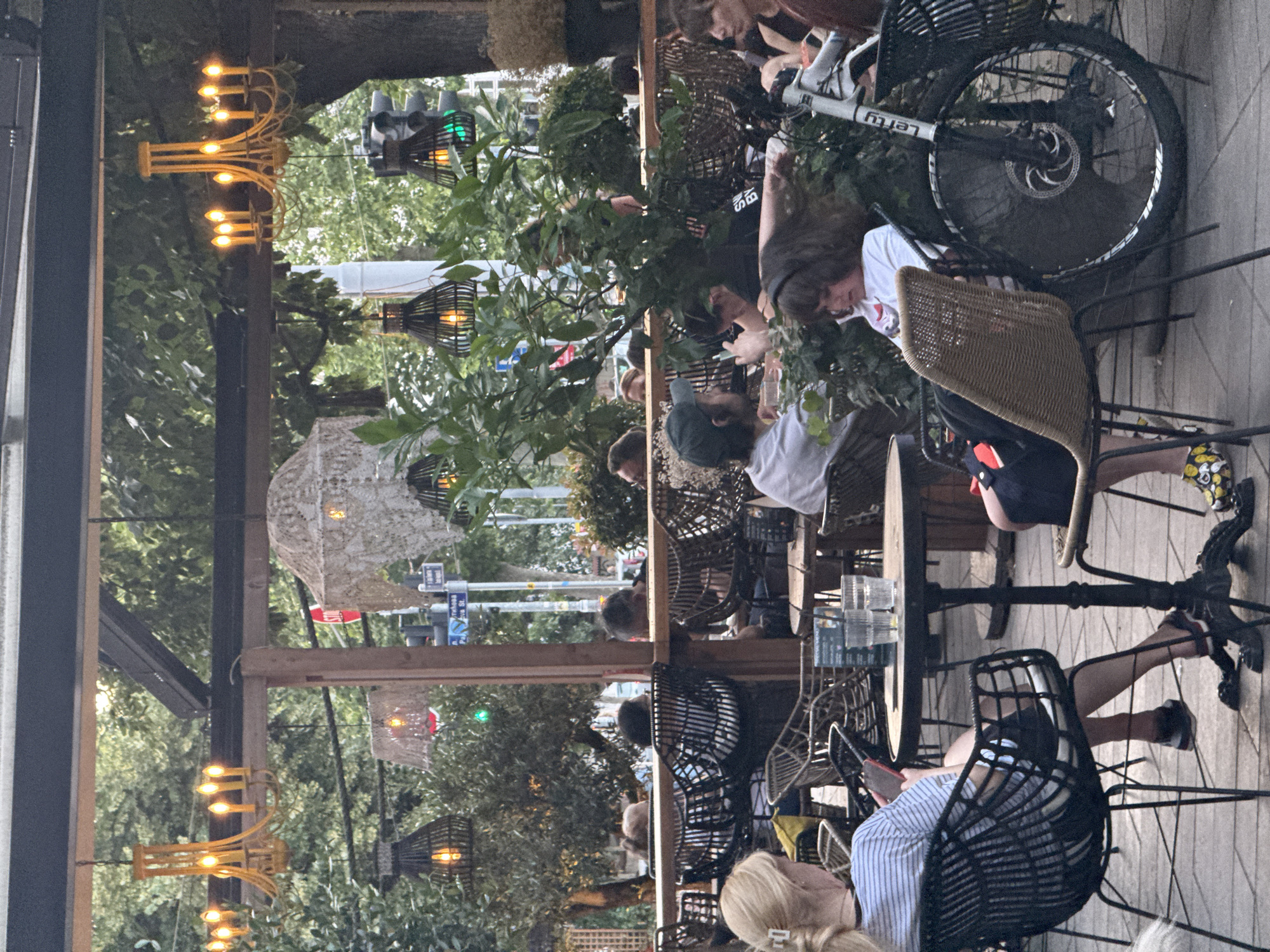 The image depicts an outdoor seating area of a cafe, likely in Belgrade, Serbia, given the street signs visible in the background. The scene is illuminated by ambient light filtering through trees and decorative pendant lights. Several patrons are seated at tables, engaged in various activities. A woman with blonde hair, wearing a striped shirt and dark shorts, is seated at a table on the left, looking at her phone. A child in a white shirt is seated at a table on the right, also looking at their phone. A bicycle with the word "Lefty" printed on the frame is parked next to the child. Other individuals are visible at other tables, some interacting with each other, others appearing to be alone. The environment suggests a relaxed, casual atmosphere. The background includes greenery, trees, and a street scene with traffic lights and street signs, confirming an urban setting. The overall impression is one of a typical cafe scene on a pleasant day.