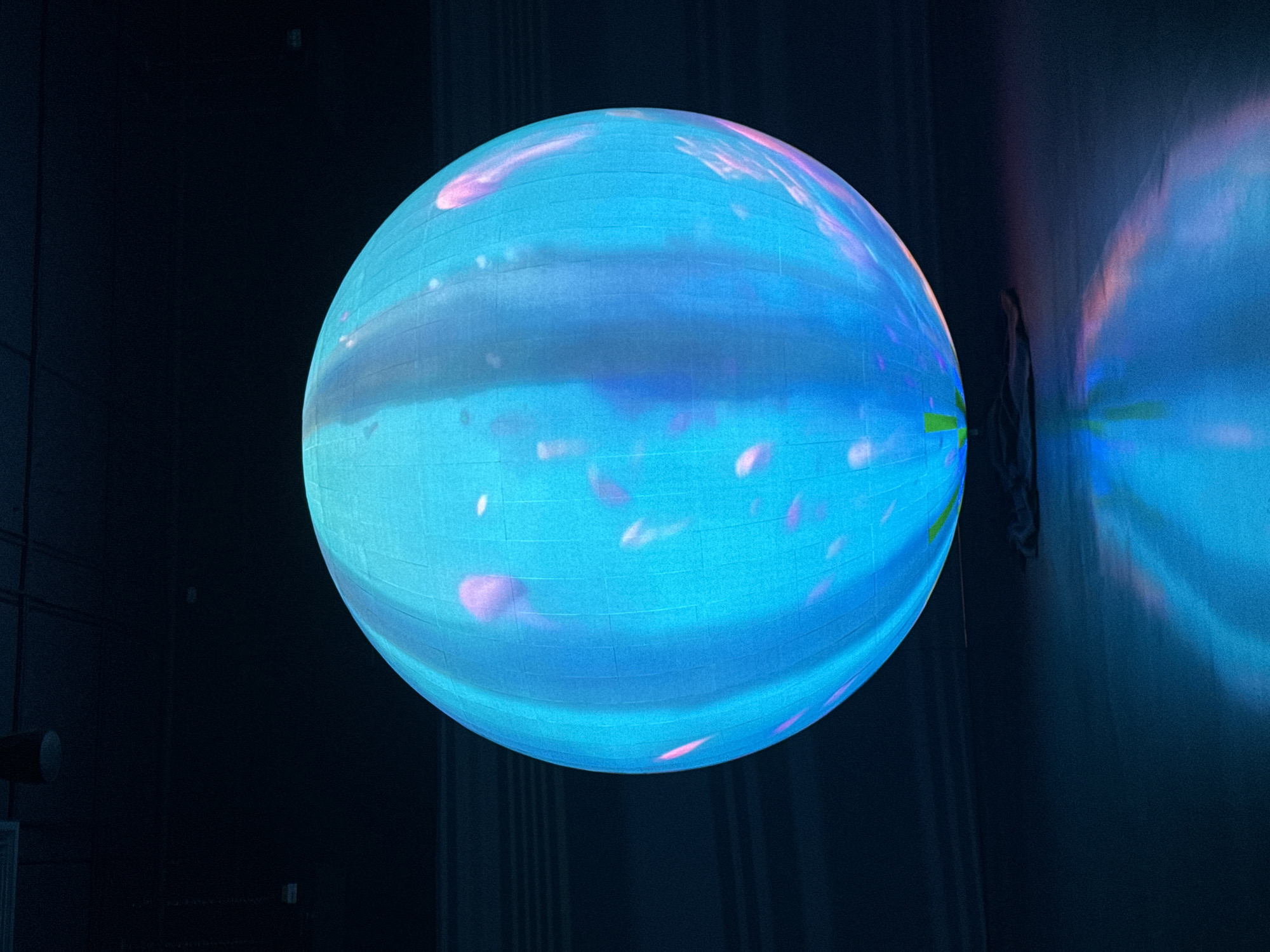 In an indoor setting with a dark ceiling and floor, a large, illuminated sphere dominates the center of the frame. The sphere, likely a projection or an LED display, emits a vibrant cyan light with subtle pink and white patterns resembling jellyfish or abstract organic shapes. The surface of the sphere appears to be segmented by vertical and horizontal lines, suggesting a grid-like structure. The sphere is resting on a dark, fabric-like material on the floor. The floor itself is dark and reflects the light from the sphere, creating a luminous pool of cyan and pink hues. The background is a dark, indistinct space, with faint horizontal lines visible on the wall to the left. The overall lighting is dim, with the sphere being the primary light source.