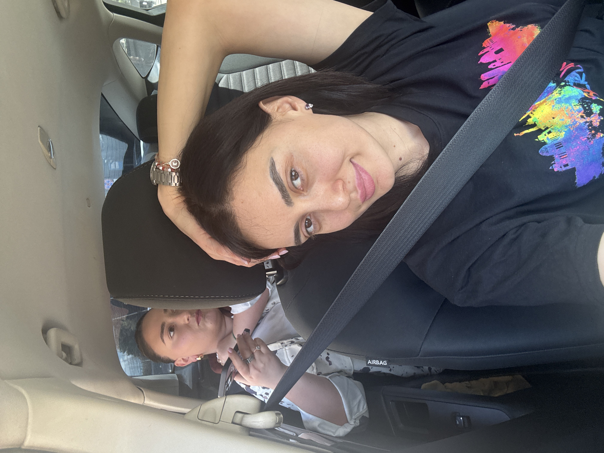 A woman with long, dark hair is sitting in the driver's seat of a car, wearing a black T-shirt with a colorful cityscape design and a seatbelt. She is leaning her head on the headrest and looking towards the camera. The car appears to be in motion based on the background scenery visible through the car window. Another woman is visible in the rearview mirror. The location is identified as Tbilisi, Georgia, based on contextual information (not visible in the image).