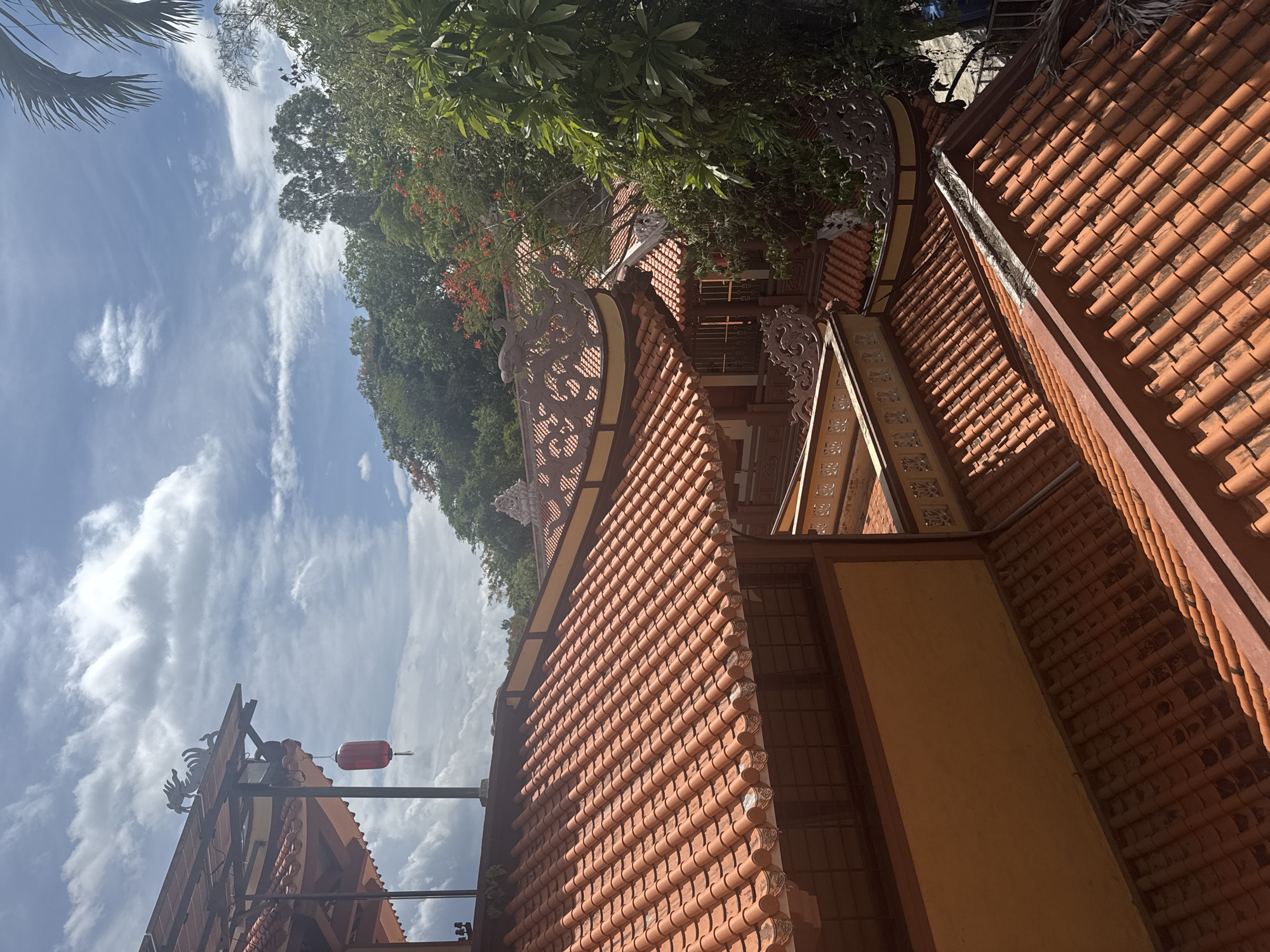 The image shows a detail of traditional Asian architecture, likely a temple or palace, with multiple terracotta tiled roofs and ornate decorative elements. The roofs are layered and have curved edges, typical of East Asian design. In the foreground, a close-up view of the tiled roof emphasizes the texture and color of the clay tiles. Further back, other sections of the building are visible, featuring yellow walls and intricate carvings along the rooflines, including what appear to be dragon motifs. The architecture is set against a backdrop of lush green trees and a blue sky with scattered white clouds, suggesting an outdoor environment in a warm climate. A red lantern hangs from one of the eaves, adding a touch of color and cultural detail. The scene is static and does not show any people or ongoing activity.