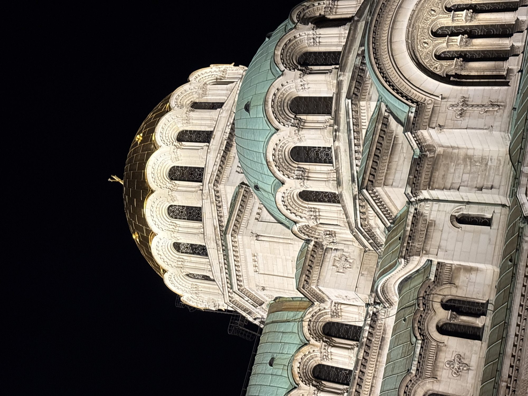 The image displays a grand architectural structure, likely a cathedral, dramatically illuminated at night. The building is presented from a low-angle perspective, rotated vertically to emphasize its towering presence against a deep black sky.
The primary subject is a multi-domed edifice, showcasing intricate and ornate architectural details. A prominent feature is a large, glowing golden dome, topped with a delicate cross, which stands out brightly. Adjacent to this, several other smaller domes and curved roof sections are covered in a distinctive greenish patina, characteristic of oxidized copper or bronze. The main body of the building is constructed from light-colored stone, adorned with elaborate carvings, columns, and decorative moldings.
Numerous arched windows, some fitted with dark grates, punctuate the facade. Intricate stonework, including relief patterns and small carved crosses, is visible, suggesting a religious building. The artificial lighting at night highlights the textures and three-dimensional elements of the architecture, creating a powerful contrast with the dark background.
The setting is outdoors, at night, with the building being the sole source of illumination in the scene. No people or specific activities are visible. Given the location context of Sofia, Bulgaria, the structure is identifiable as the Alexander Nevsky Cathedral, renowned for its iconic golden and green domes and ornate Neo-Byzantine design, especially when lit up after dark. There is no visible text in the image.