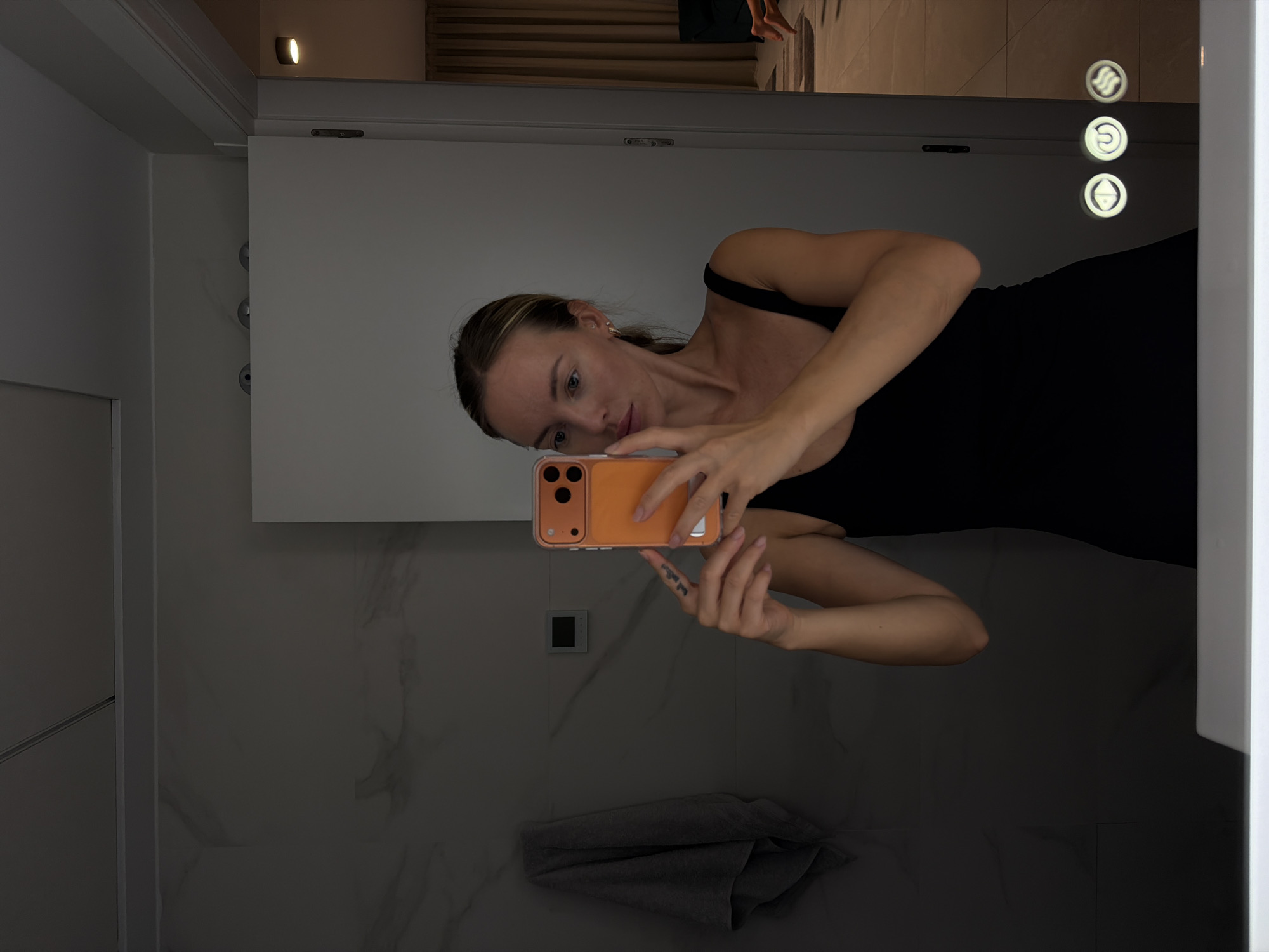 A woman with blonde hair pulled back in a ponytail is standing in a modern bathroom and taking a mirror selfie. She is wearing a black tank top and holding an orange smartphone in her right hand. The bathroom has marble-patterned walls, a towel hanging on the left, and a light-up mirror with three circular icons on the bottom right. In the reflection of the mirror, another person can be seen in the background, possibly in an adjacent room or hallway. The overall aesthetic suggests a clean and minimalist design. There is no indication that this location is in Finestrat, Spain.