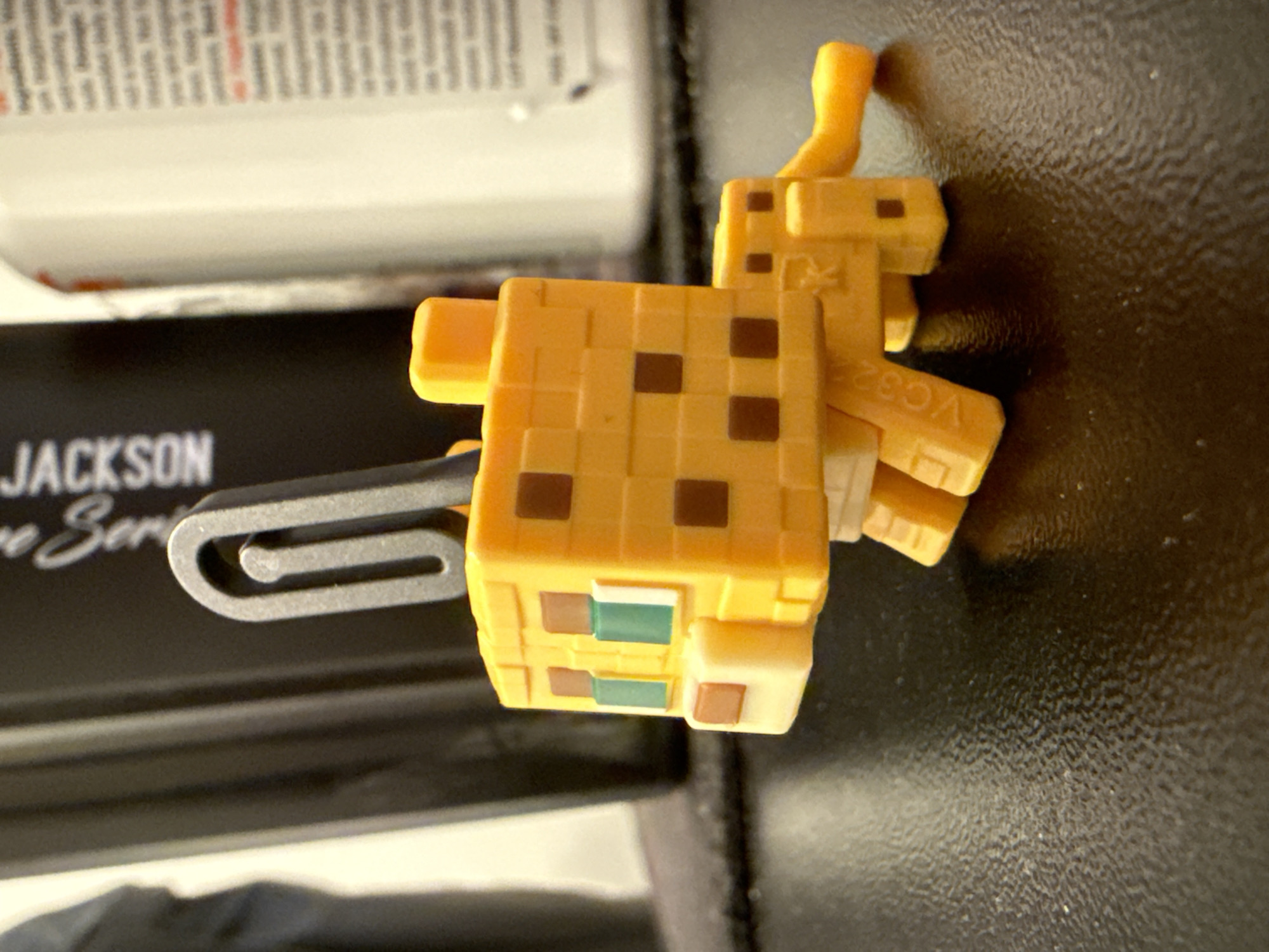 The image displays a small, blocky, yellow-orange toy resembling a Minecraft ocelot or cat, positioned horizontally on a dark, textured surface. The toy features dark brown square spots across its body, with additional green and light brown/white block details on its face. It has small, square ears and a short, curled tail. A silver-colored metal clip or hook is attached to the toy's left side, indicating it may be a keychain or similar accessory.
In the out-of-focus background, a white object with fine, unreadable print is visible at the top. Below this, a black object, likely a book or case, shows partial white text reading "JACKSON" and "ve Ser." The dark surface beneath the toy has a subtle, uneven texture, possibly a fabric or artificial leather. Visible text on the toy includes "VC32" stamped on its back leg, and a small, stylized symbol that appears to be a 'K' or star near its shoulder. The scene is lit from an overhead source, casting a soft shadow from the toy onto the textured surface. The toy is stationary, with no specific activity taking place.