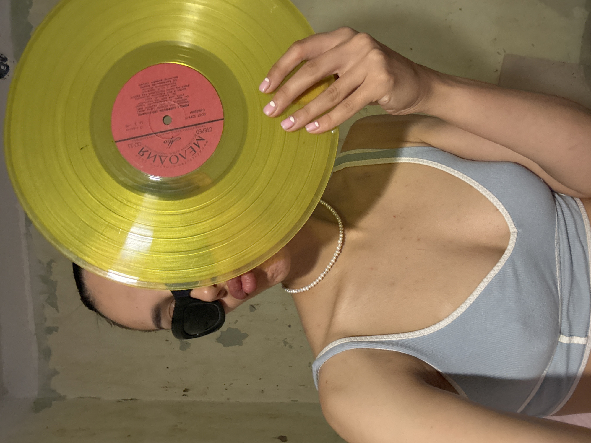 A young woman is shown from the chest up, holding a yellow vinyl record in front of her face. She is wearing a light blue ribbed tank top with white trim and a pearl necklace. Her lips are pursed, and she is wearing black sunglasses. The background appears to be a wall with peeling paint. The vinyl record has a pink label with Russian text. This image was captured inside a building, likely a residential space, and there is no indication of its specific location within Votkinsk, Russia, or any outdoor elements.
