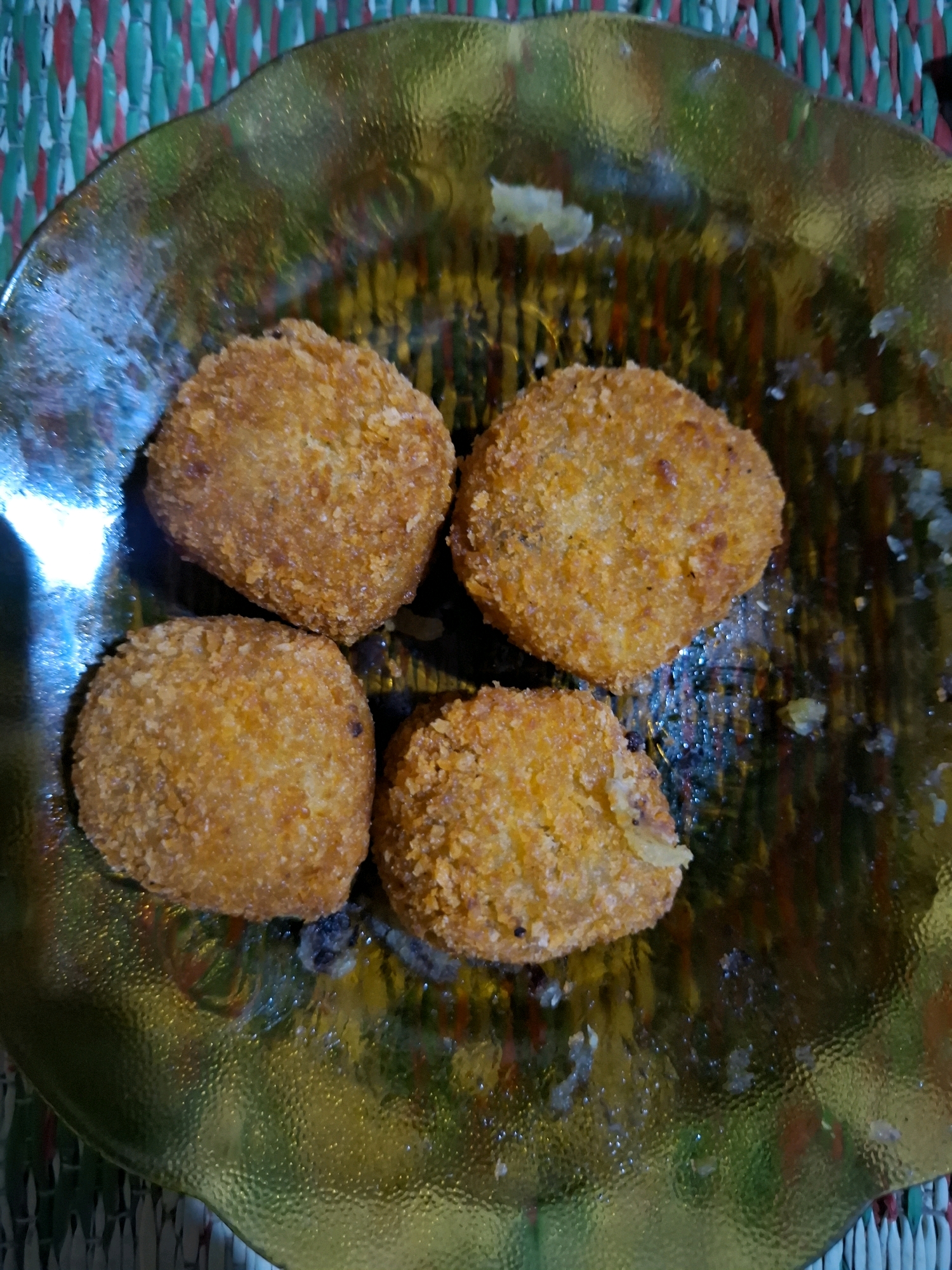 The image shows four golden-brown, breaded croquettes or nuggets arranged on a textured, translucent green glass plate. The plate is set on a woven mat with alternating red and green threads. The croquettes are roughly oval in shape and appear to be fried, with a crispy exterior. Some small crumbs and dark specks are visible on the plate around the croquettes. The lighting creates a reflection on the surface of the plate, highlighting its textured pattern. The location is identified as Sembung, Indonesia.