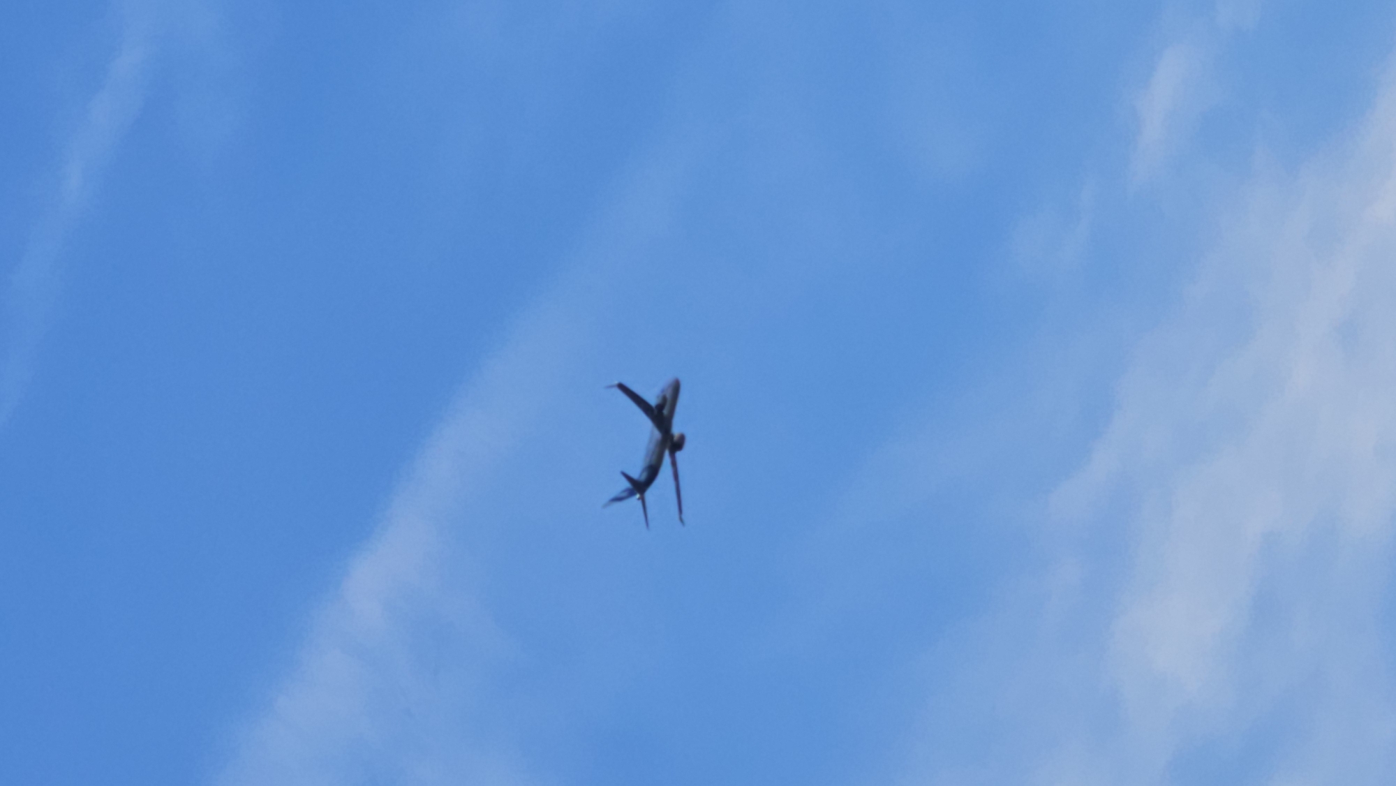 The image shows a passenger airplane in the sky. The airplane is viewed from below, and it appears to be in the process of banking or turning. The sky is a clear blue, with some scattered white clouds. The lighting suggests it is daytime, possibly mid-morning or afternoon. The image is taken from the ground, looking upwards. There are no people visible, only the airplane as the primary object. The setting is an open sky, indicative of a flight path over an outdoor area. The scene is peaceful and ordinary, depicting a common occurrence of air travel. There is no visible text in the image. The location context provided is Панфилово, Kazakhstan, which implies the airplane is flying over this region.