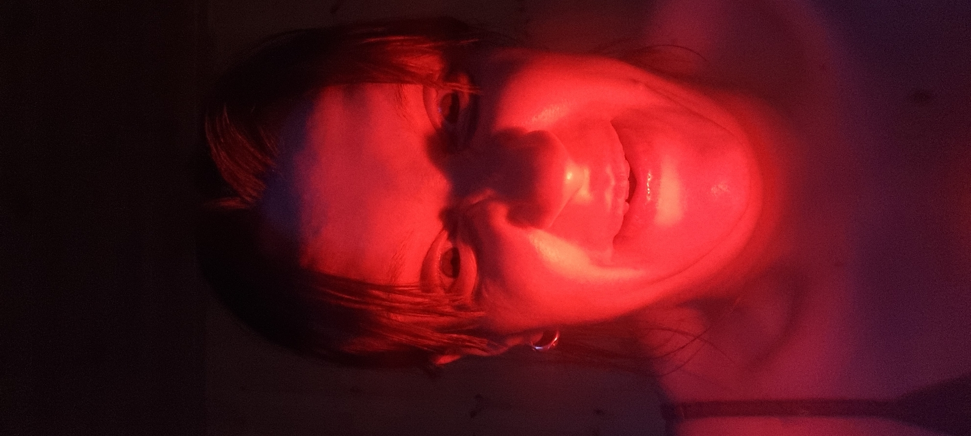 A close-up, horizontally oriented shot reveals a person's face illuminated by red light. The person has dark hair and their head is tilted, appearing as if they are lying down or leaning back. The surrounding environment is dark, suggesting a dimly lit space. No specific details indicate a location in Dunnae-myeon, South Korea.