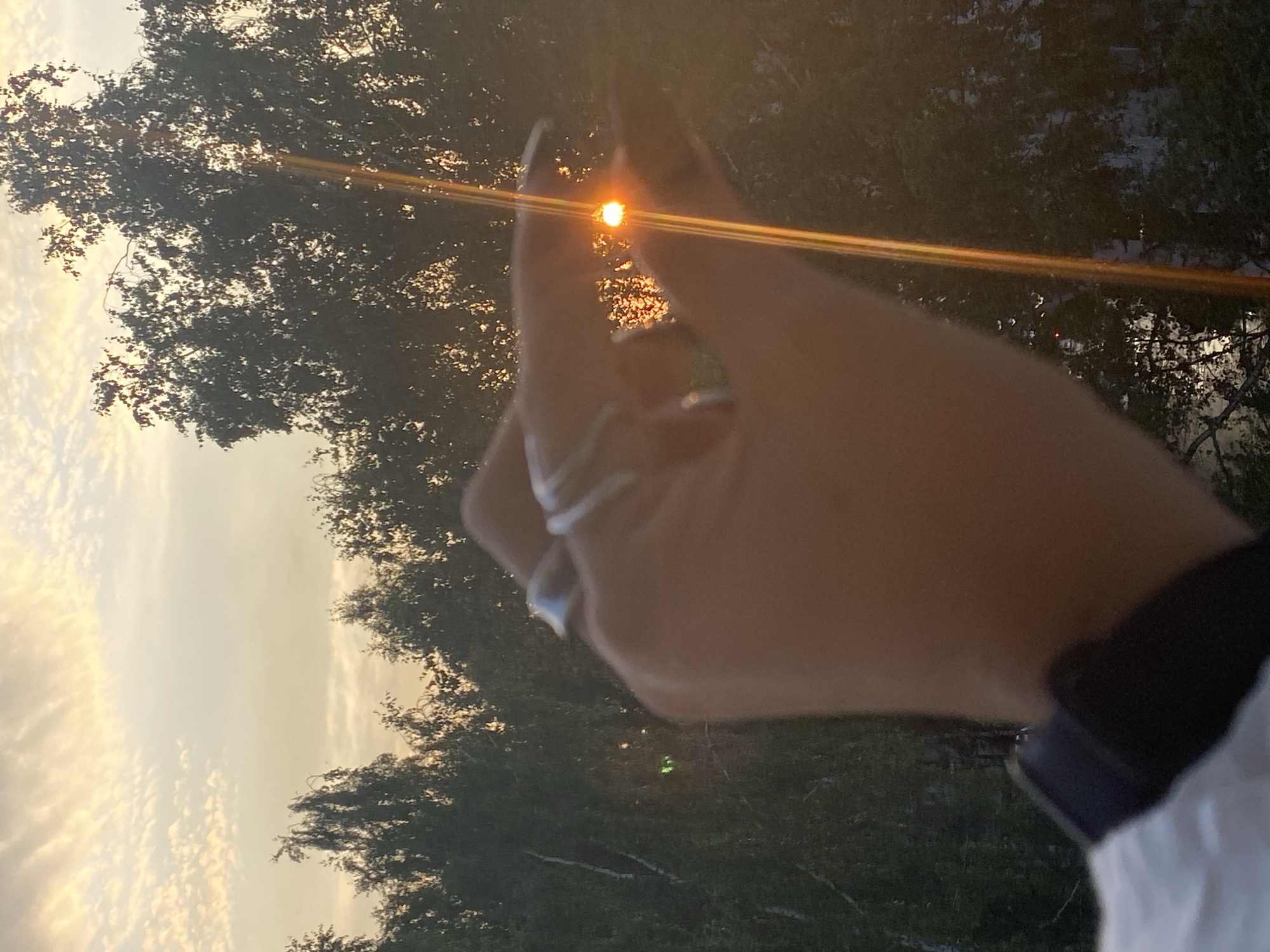 A hand wearing rings is held up against a sunset sky, with the sun's rays filtering through the fingers. The hand is positioned in the foreground, with a blurred background of trees. The sky is a soft orange and pink, with scattered clouds. The sun is a bright, small orb, creating a lens flare effect. The overall mood is peaceful and serene. The location is Chaikovsky, Russia.