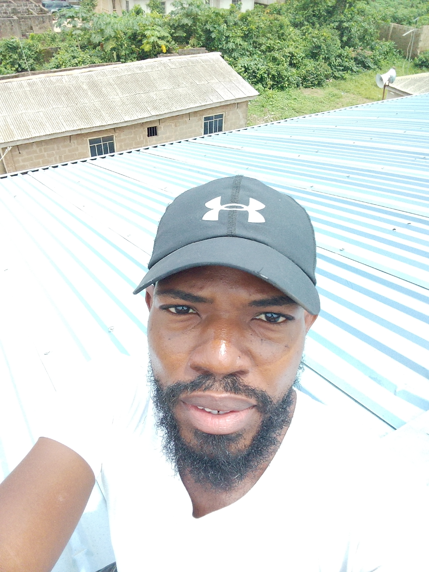 A selfie captures a man wearing a black Under Armour baseball cap and a white t-shirt. He has a beard and is looking at the camera. The background shows a corrugated metal roof with blue and white stripes, and in the distance, there are buildings and lush green vegetation, suggesting an outdoor urban setting. This scene is likely located in Agege, Nigeria, a densely populated area known for its diverse architecture and vibrant natural elements.