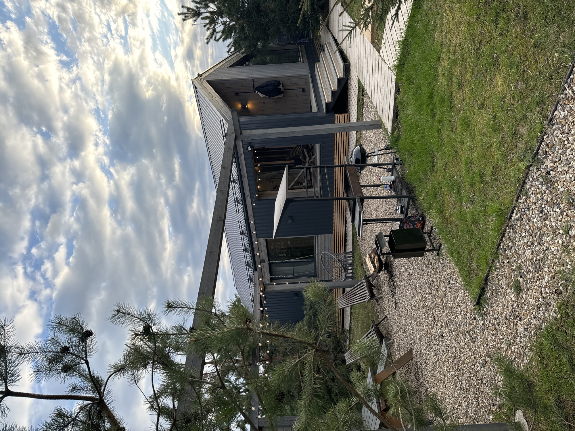 The image shows the exterior of a modern, dark-colored wooden house with an A-frame roof and a wooden walkway leading to the entrance. A small yard with grass and gravel surrounds the house. There is outdoor furniture including chairs, a grill, and a fire pit. String lights are hung along the eaves of the house. A cloudy sky is visible above. The location is Литвиново, Russia.