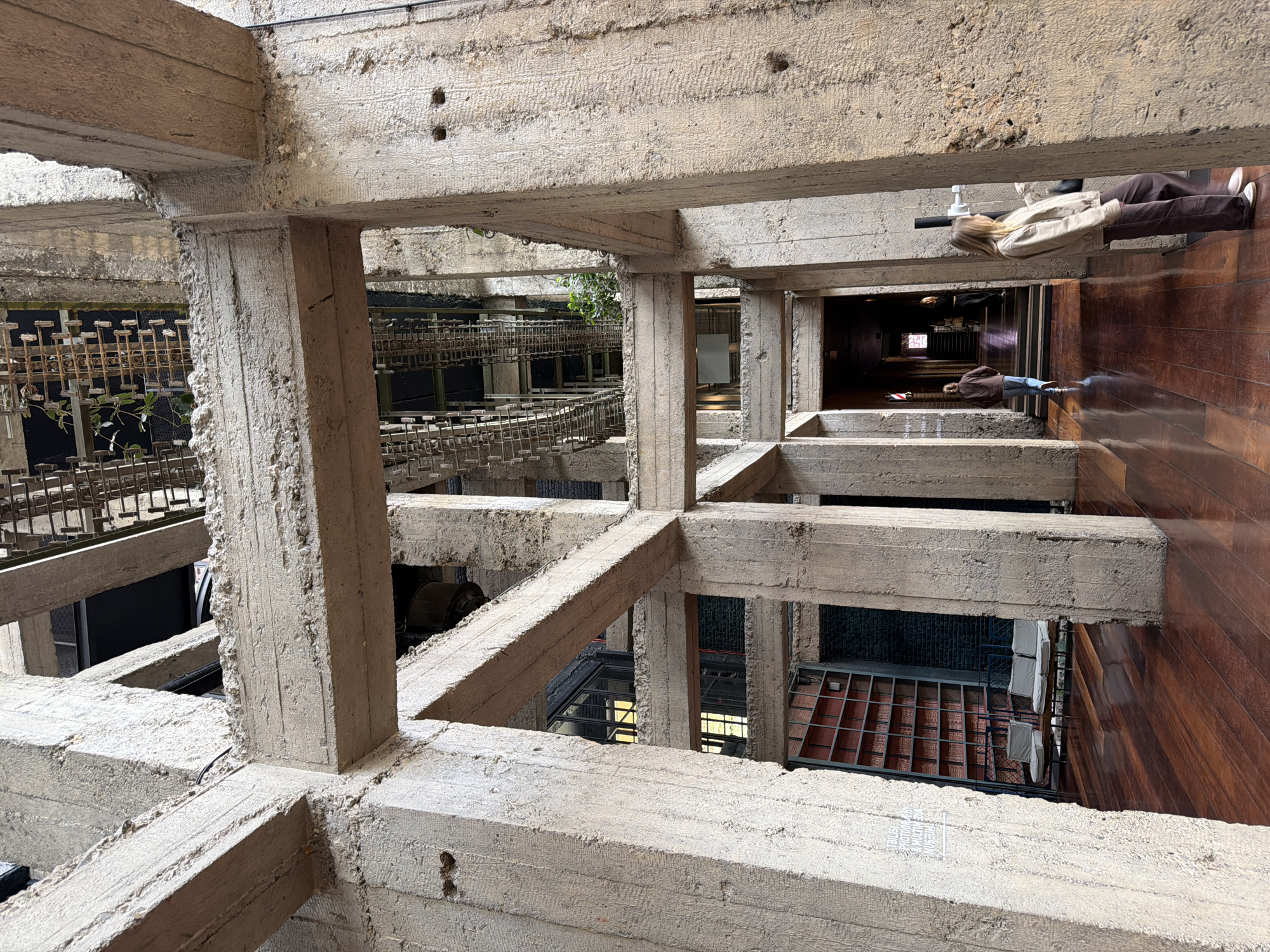 The image shows an interior space with a grid of concrete beams and pillars forming a structural framework. This is likely the Fabrika Hostel and Suites in Tbilisi, Georgia. The structure creates a series of rectangular openings offering views to other areas.
To the right, a polished wooden floor is visible along with people walking. One individual is wearing brown pants, white shoes and a light jacket. Further down, there are more people visible in a hallway.
To the left, there is a metal structure, possibly a staircase or walkway, with plants visible in the background.
The concrete structure itself is rough in texture, with visible markings and imperfections. The lighting appears to be a mix of natural and artificial.