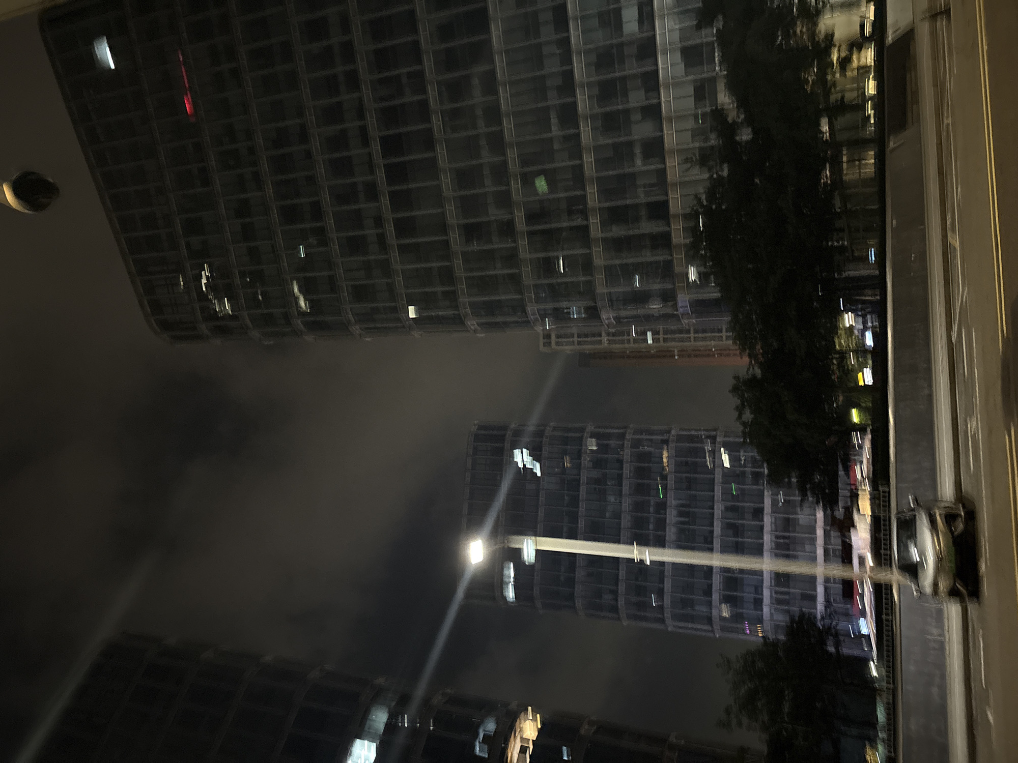 This is a nighttime photograph, taken from a low angle, looking up at several modern, glass-facade skyscrapers. The buildings are brightly lit from within, with many windows showing a grid of lights. The sky is dark and cloudy, with a soft glow suggesting artificial lighting from the city.
In the foreground and middle ground, a street lamp with a bright white light extends horizontally across the image, casting a beam that reflects on the wet street. A dark car is visible on the right side of the street, its headlights on. Trees with dark foliage are interspersed between the buildings, partially obscuring the view of the lower floors.
The overall impression is one of a bustling urban environment at night. The scene appears to be a city street in Haizhu District, China, characterized by contemporary architecture and urban infrastructure. There are no visible people or text in the image. The weather appears to be overcast or foggy, contributing to the diffused lighting and slightly hazy atmosphere. The mood is one of quiet, nocturnal activity.