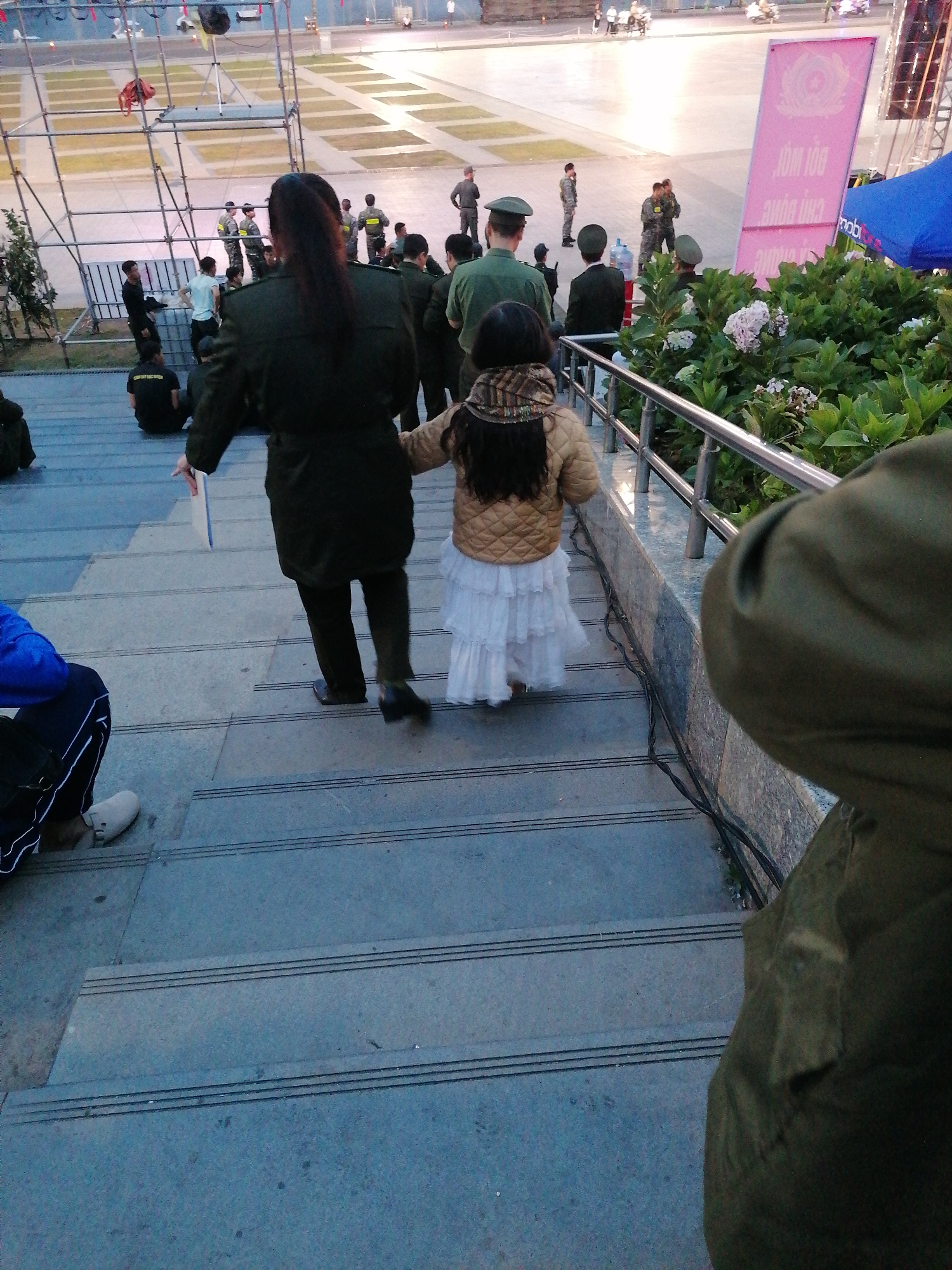 The image shows a group of people on a set of outdoor stairs. In the foreground, a person in a dark uniform is walking up the stairs, holding a white object. Beside them, a young girl in a light-colored quilted jacket and a white, tiered skirt is also walking up the stairs. In the background, several individuals in military-style uniforms are visible, some standing and some walking.
The scene appears to be an outdoor public space, possibly a plaza or an event venue, with tiered seating or steps. Behind the people on the stairs, there are metal structures and fencing, suggesting a temporary setup for an event. Further in the background, there are more people, some in uniforms and some in civilian clothing, and what looks like a stage or platform. A large, pink banner with text is visible on the right side of the image. The lighting suggests it is either early morning or late afternoon, with a soft, diffused light.
The overall atmosphere seems orderly, with people moving purposefully. The presence of uniforms and the structured environment might indicate an official gathering, a parade, or a ceremony. The people on the stairs are the central focus, with the woman in uniform and the young girl being the most prominent. The image is taken from a slightly elevated perspective, looking down on the subjects as they ascend the stairs.