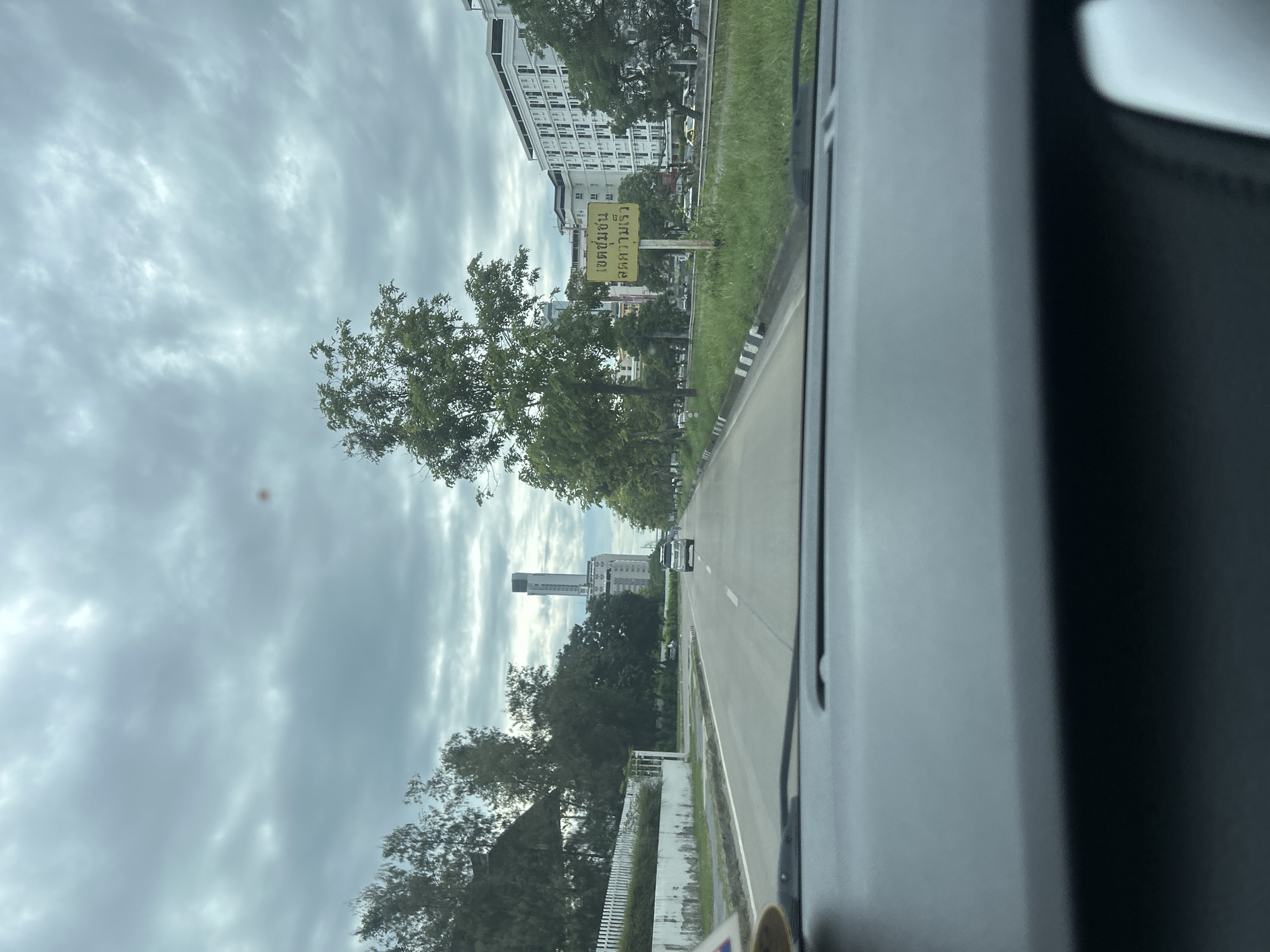 The image is taken from inside a vehicle, looking out at a road in Tha Khlong Town Municipality, Thailand. The road is paved and has multiple lanes, with white dashed lines marking the lanes. A single silver SUV is driving in the right lane. On the left side of the road, there is a white fence and lush green trees. On the right side of the road, there is a grassy median with a yellow sign displaying Thai text. Beyond the median, there are several trees and modern buildings, including a tall, slender skyscraper in the distance. The sky is overcast with thick, gray clouds. The foreground shows the dark dashboard of the vehicle.