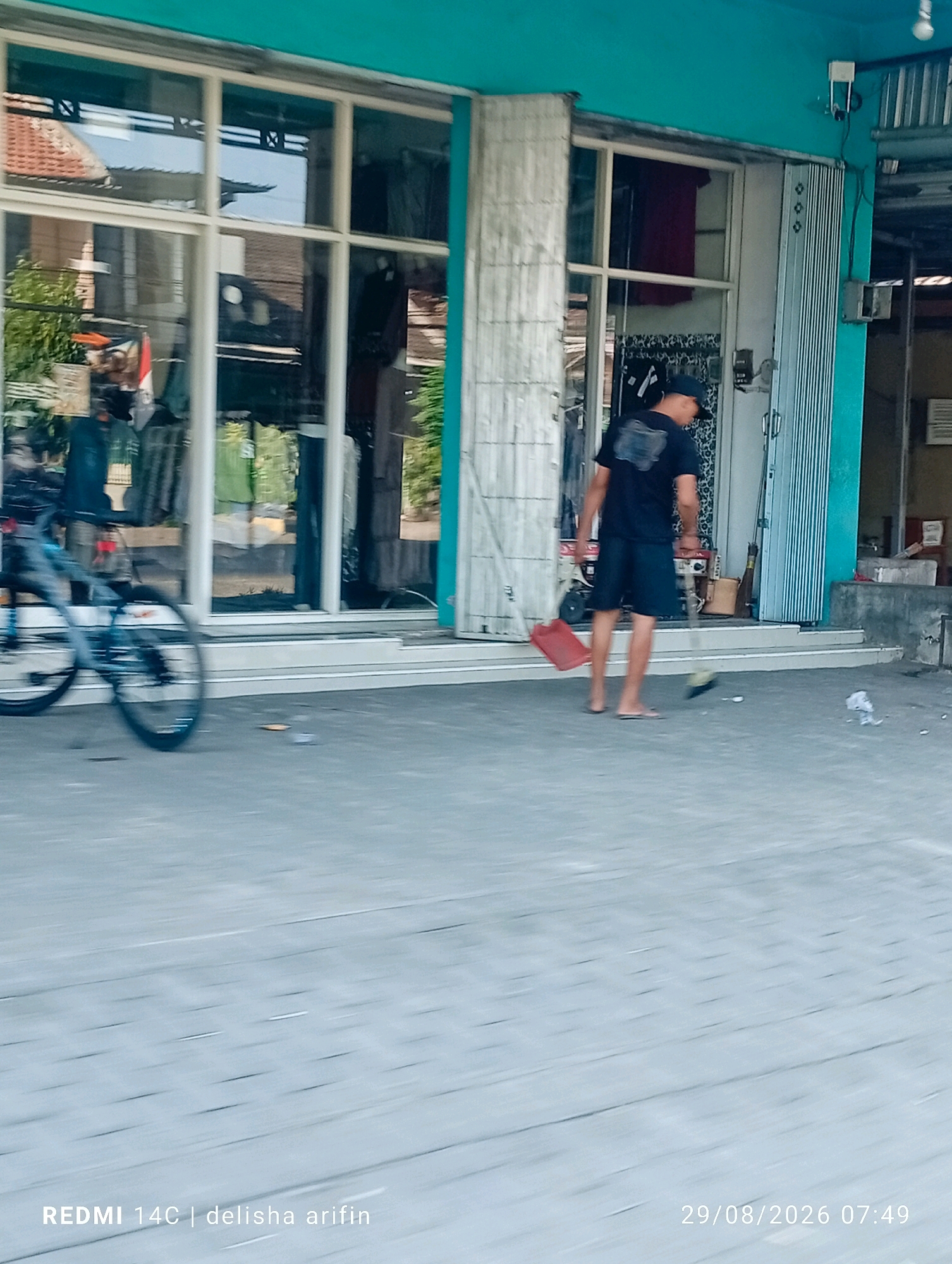 In Wonorejo, Indonesia, a man wearing a black t-shirt and shorts is sweeping the sidewalk in front of a shop. He is using a broom and a red dustpan. A bicycle is parked to the left of the scene. The shop has a teal-colored facade and large windows displaying merchandise. The sidewalk is paved with gray tiles. The image is dated 29/08/2026 at 07:49.