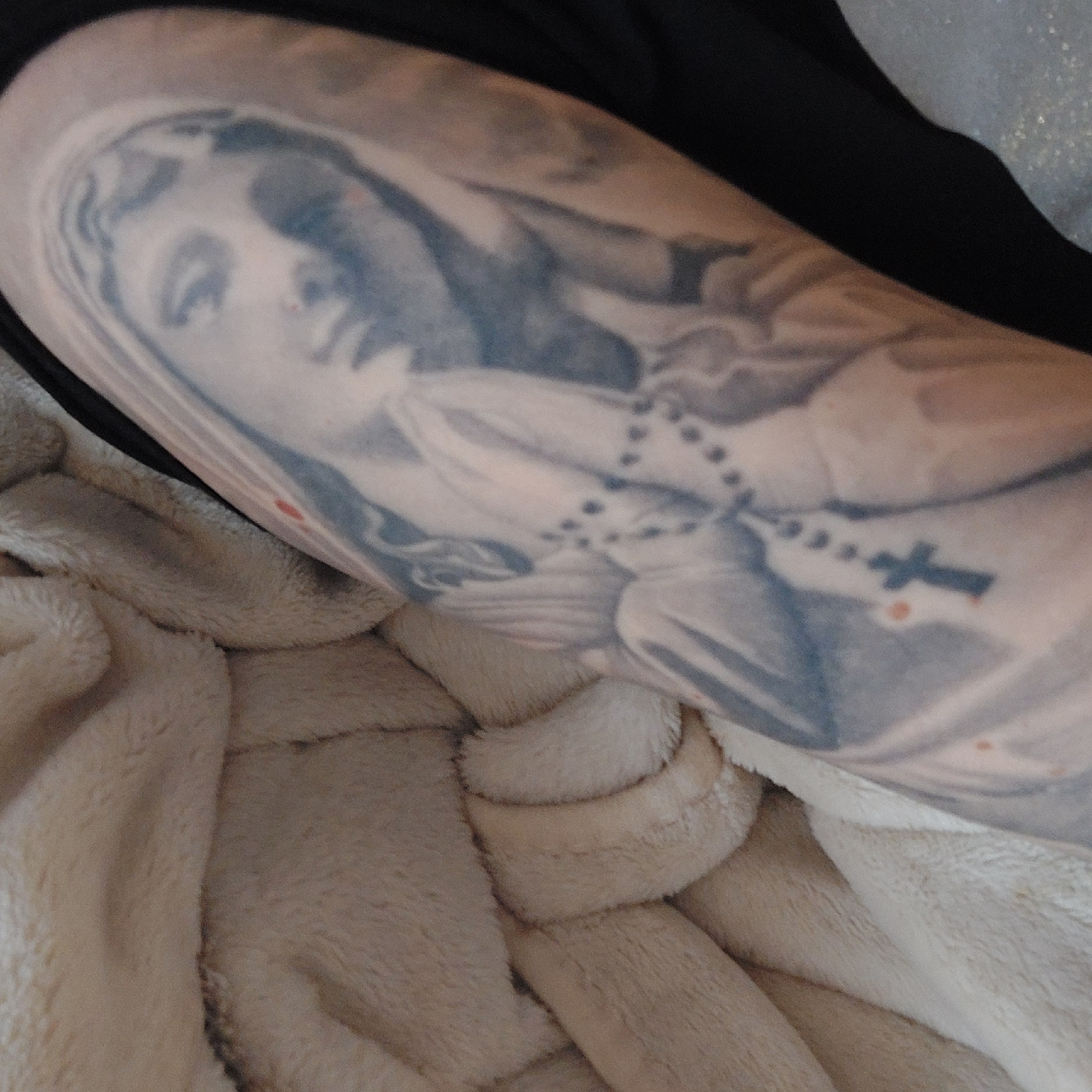 A close-up shot depicts a person's arm, bearing a tattoo of the Virgin Mary with a rosary. The background includes a soft, beige, textured blanket and a portion of a black garment. There is no information in the image that can be used to connect the scene to the city of Plovdiv, Bulgaria.