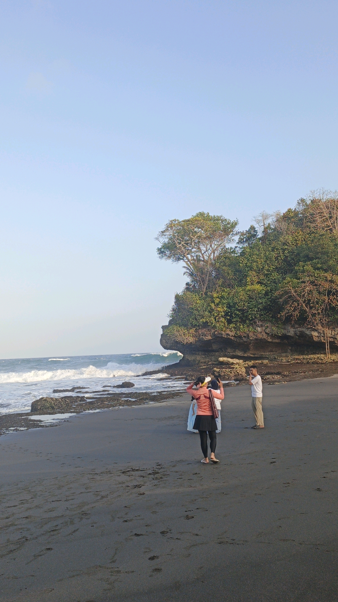 A full shot captures three people standing on a dark sandy beach, facing the ocean under a clear, pale blue sky. A large, rocky cliff covered in lush green trees dominates the right side of the frame, with waves crashing against its base. The person in the foreground, wearing a coral-colored jacket, black leggings, and a white visor, has their hands on their head and is looking down. To their left, a person in a white dress stands partially obscured, and to the right, a man in a white t-shirt and tan pants is seen from the chest up, holding his hand to his face as if shielding his eyes. The beach is scattered with rocks and debris, and the ocean stretches to the horizon.